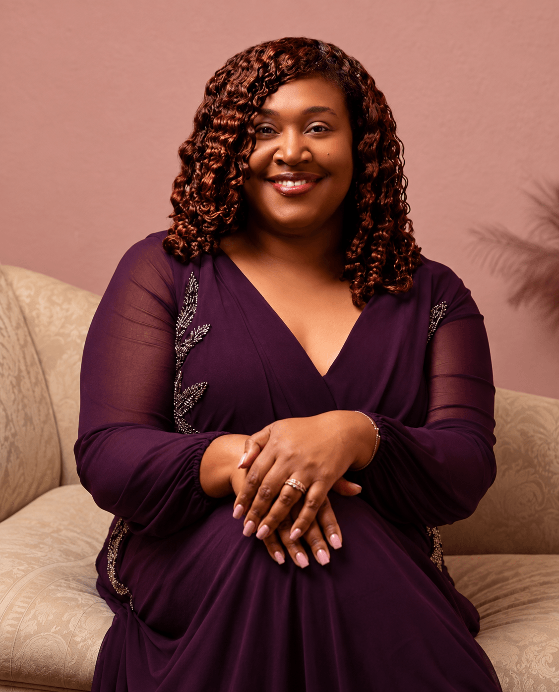 Smiling woman with curly hair sitting on a beige couch, wearing a dark purple dress, hands clasped, pink background.