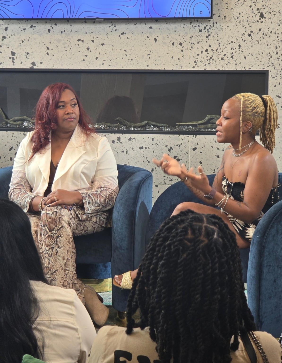 Two women seated and talking, one with curly hair and the other with braided hair, in a room with a concrete wall.
