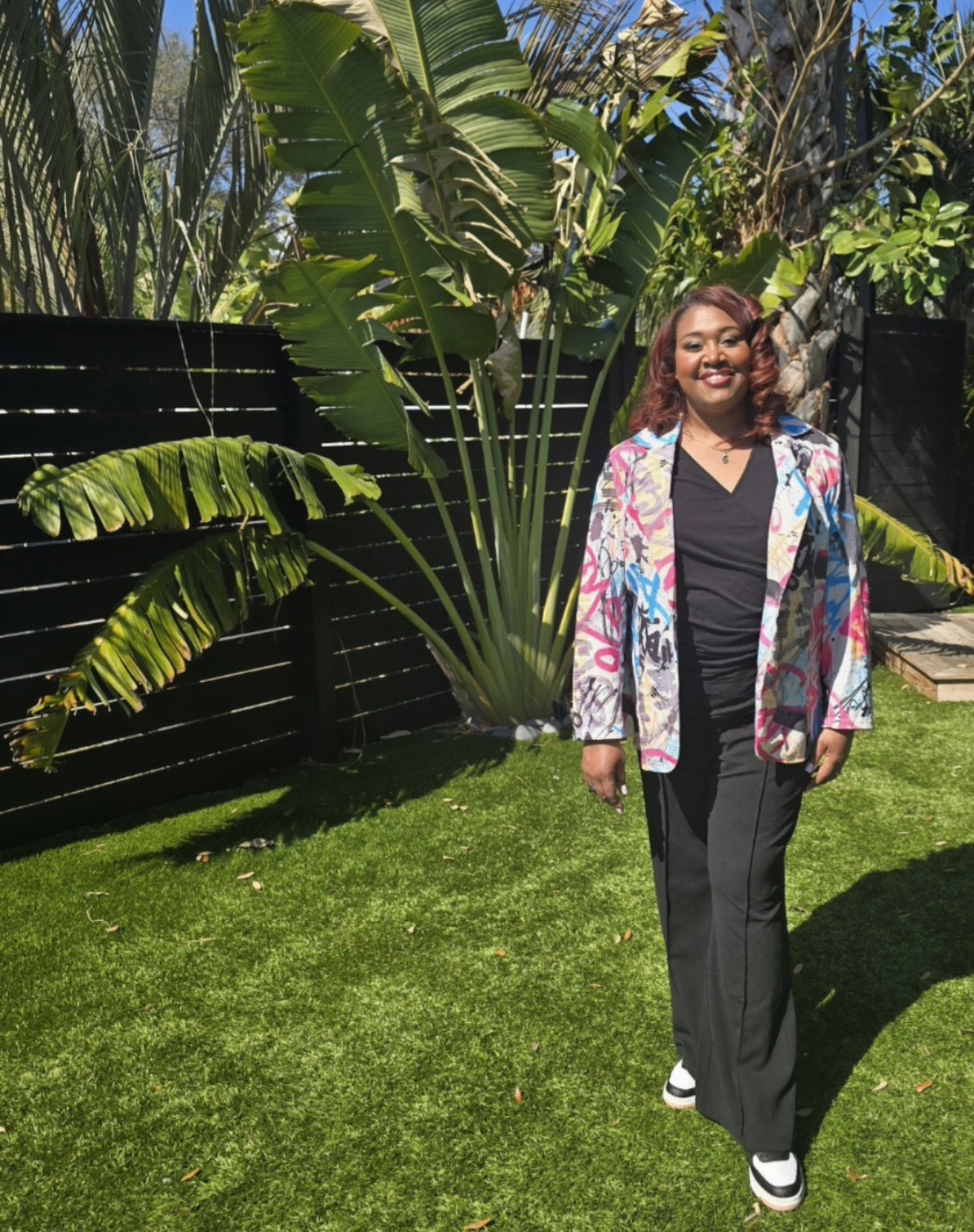 Woman with curly hair smiling outdoors, standing on grass with large green plants and a black fence behind her.