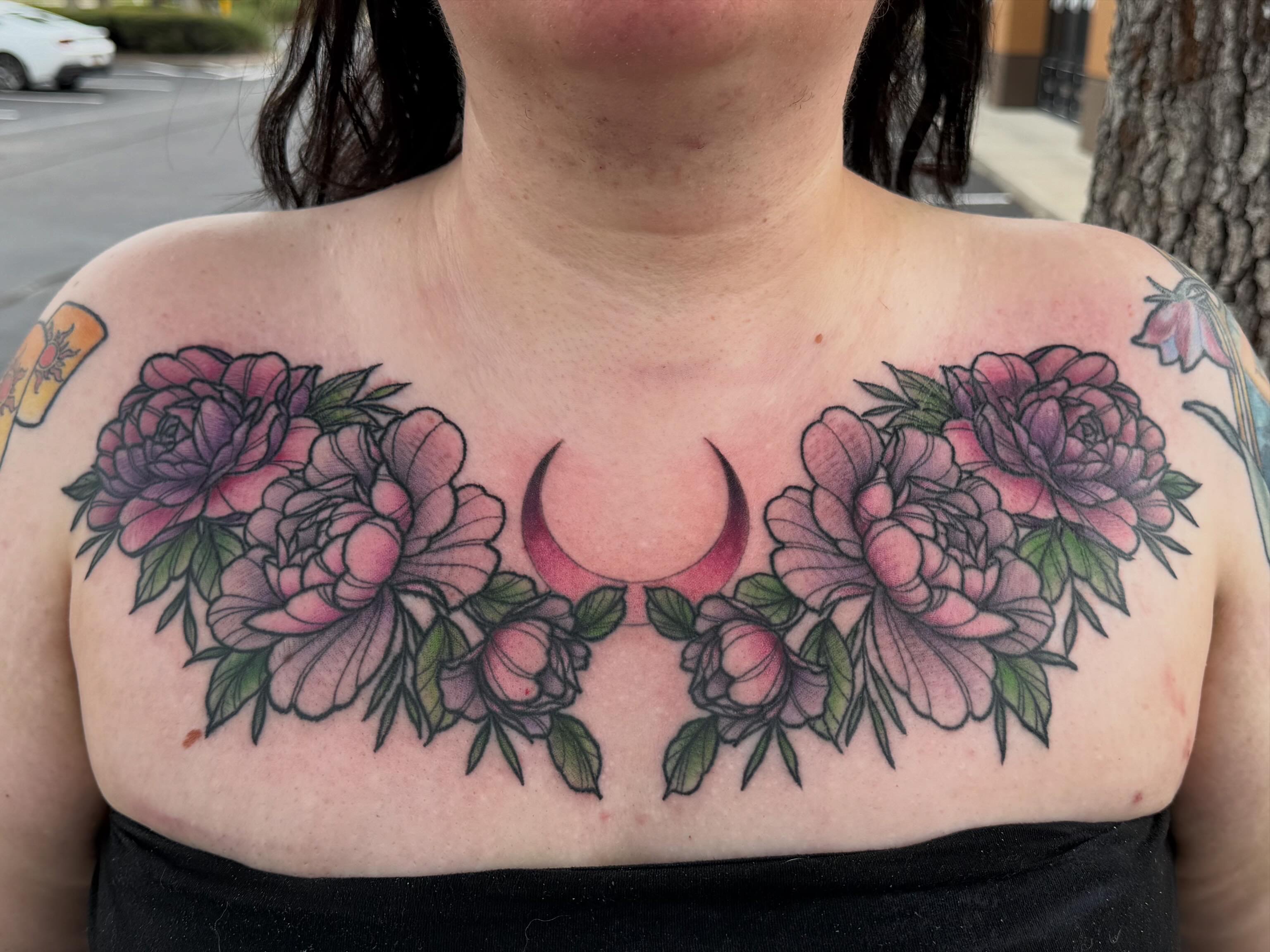 Tattoo of pink peonies and green leaves across the chest with a crescent moon in the center.