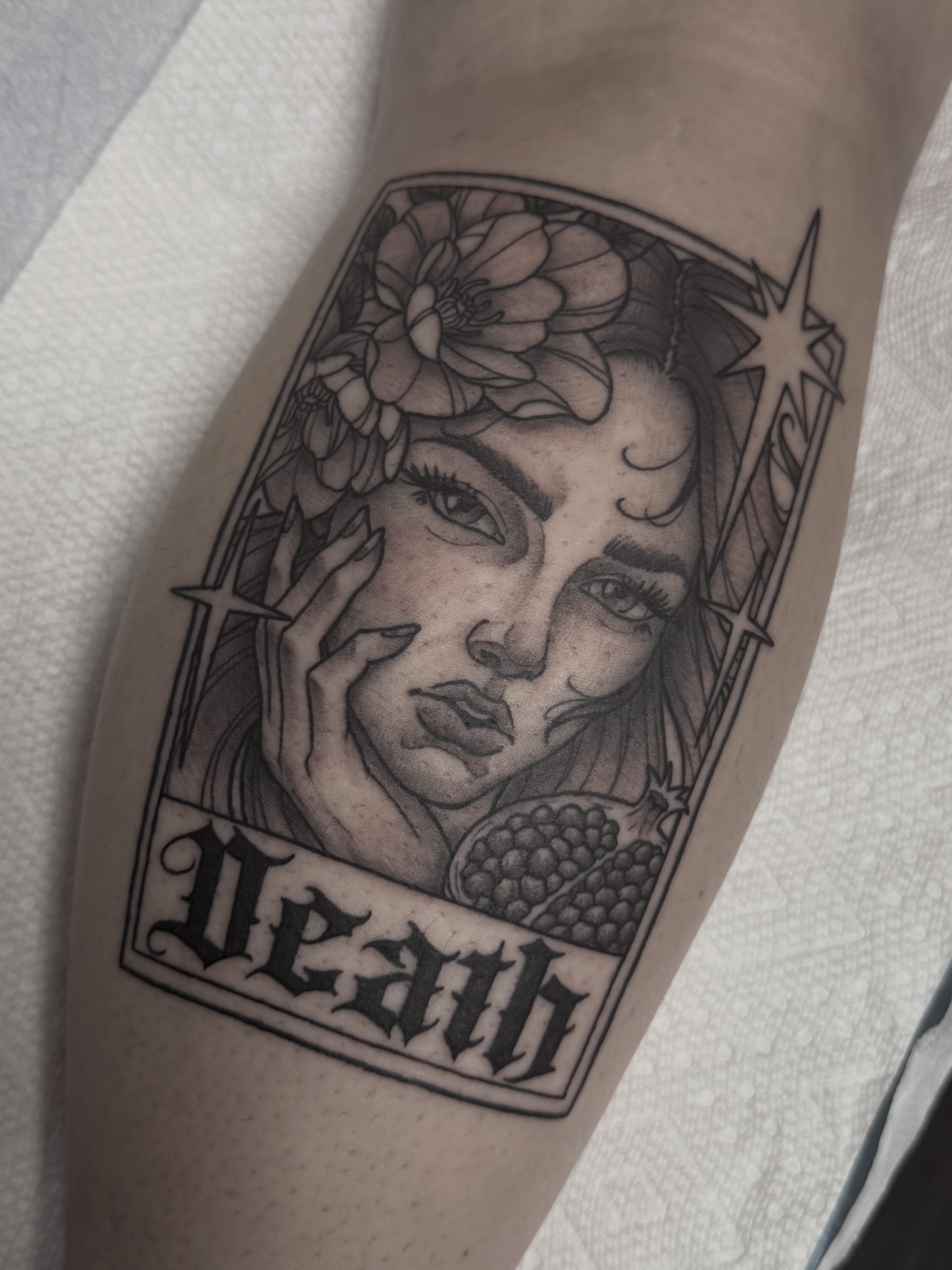 Black and gray tattoo of a woman's face with flowers, stars, and the word 'Death' below, on a person's arm.