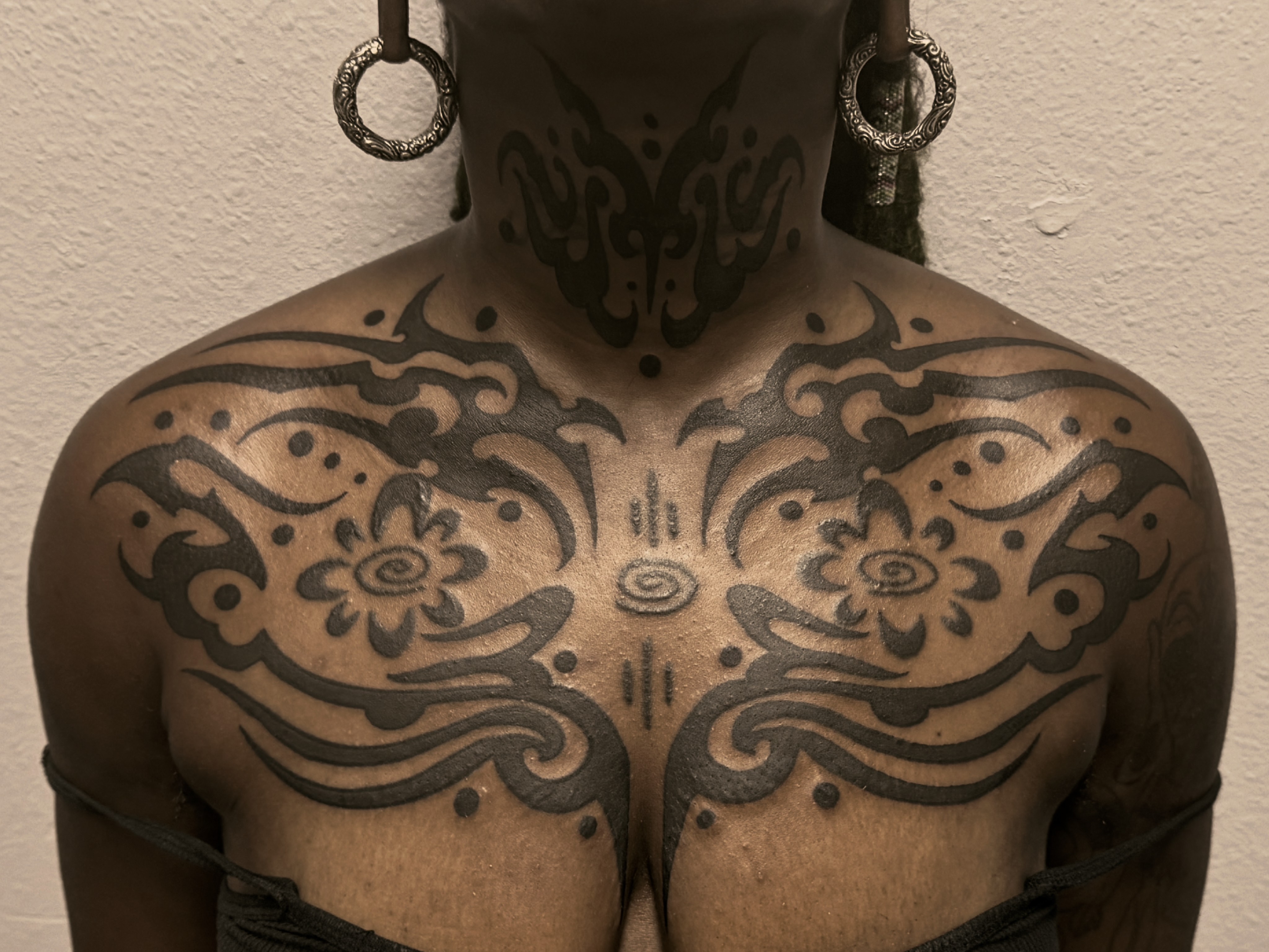 Person with large black tattoos on chest and neck, earrings, and textured wall background.