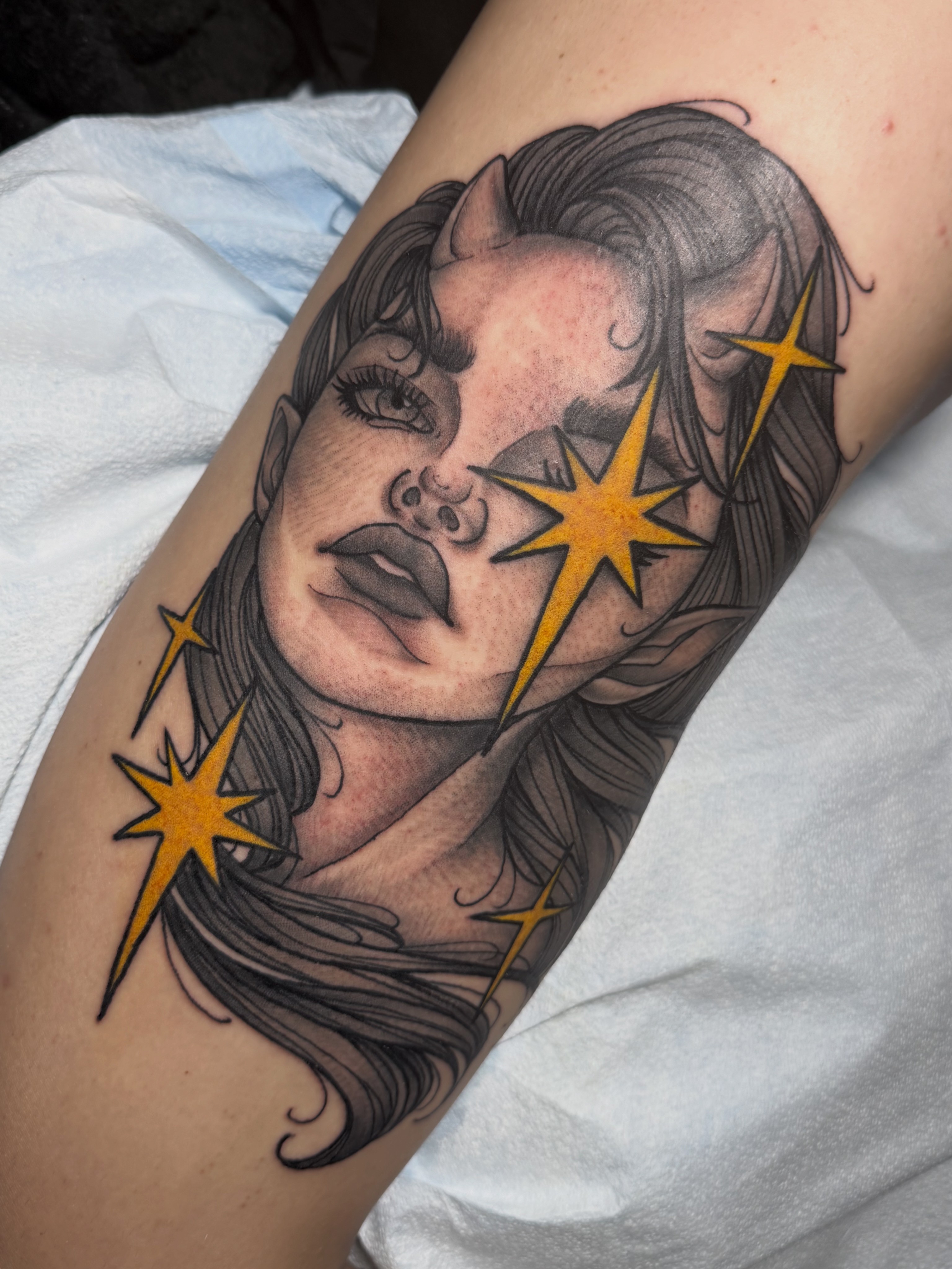 Black and gray portrait of a woman with wavy hair, surrounded by yellow starburst shapes, on a person's arm.
