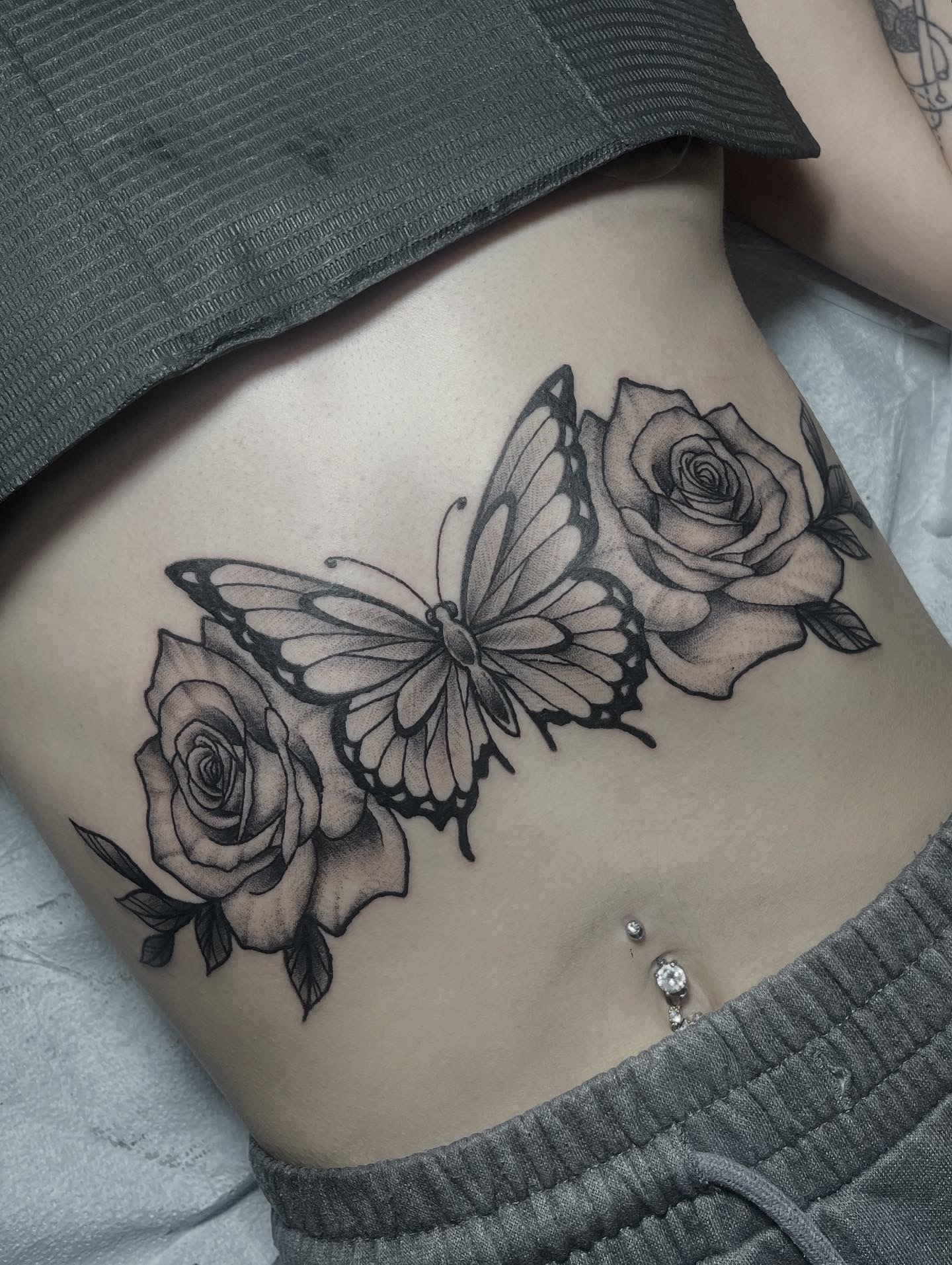 Tattoo of two roses and a butterfly across the lower abdomen, with a belly button piercing below.