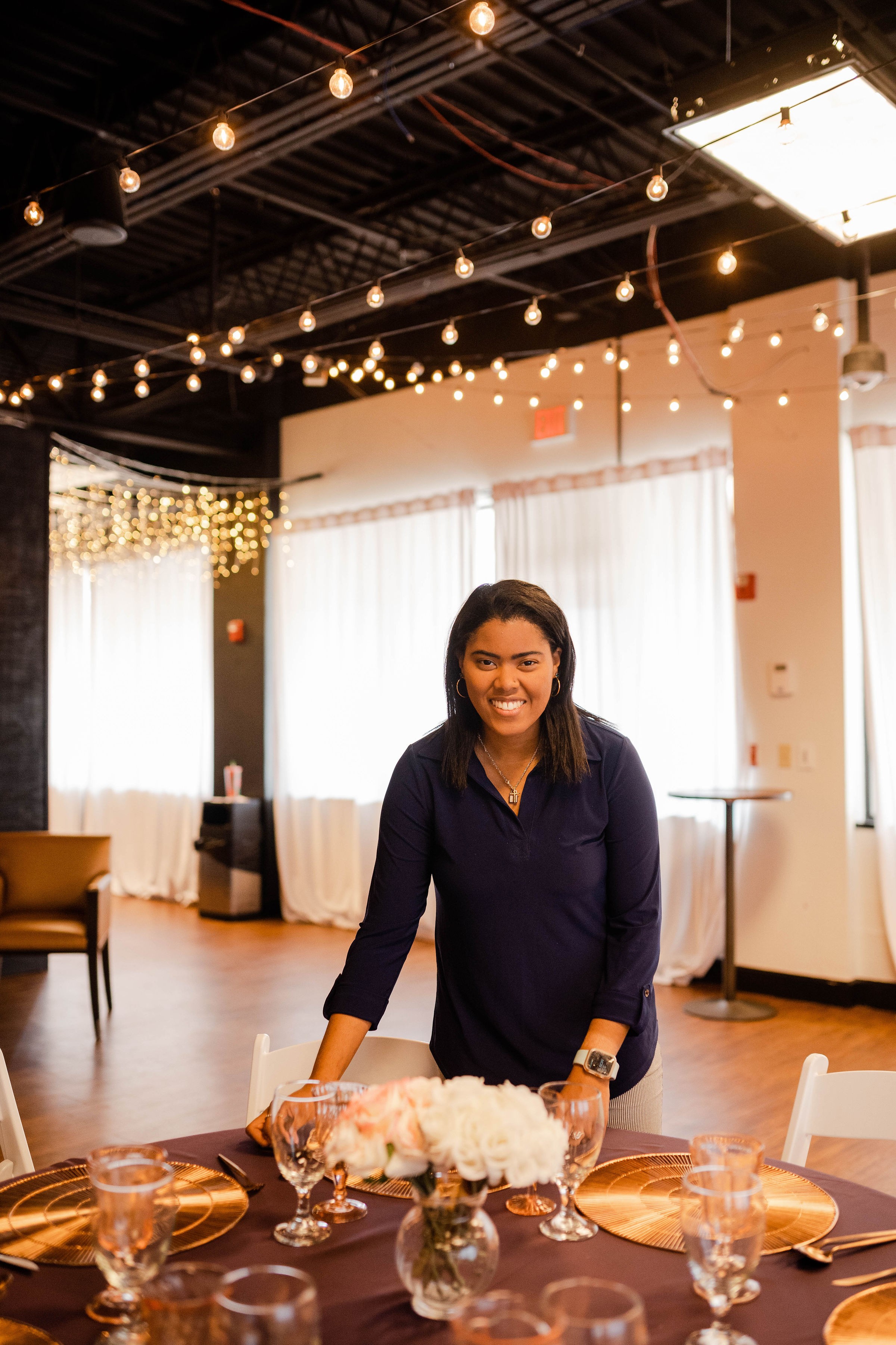 Woman standing behind a table with a cake and glasses, decorated for an event, in a room with string lights and curtains.