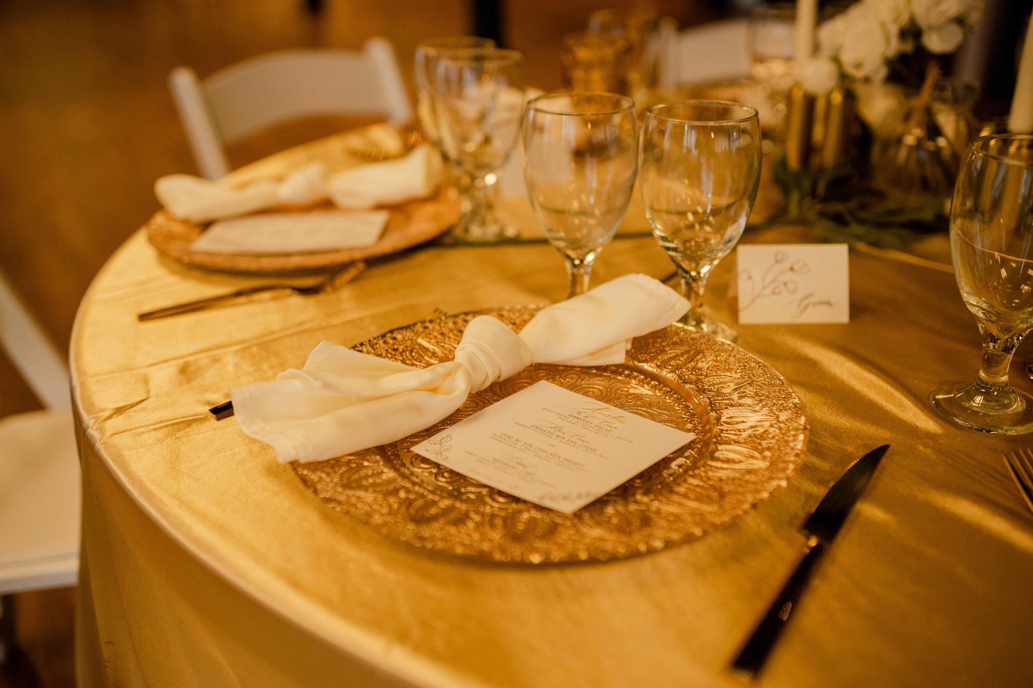 Round table set with glassware, napkins, and place cards, with a gold-colored placemat and a menu card, in warm lighting.