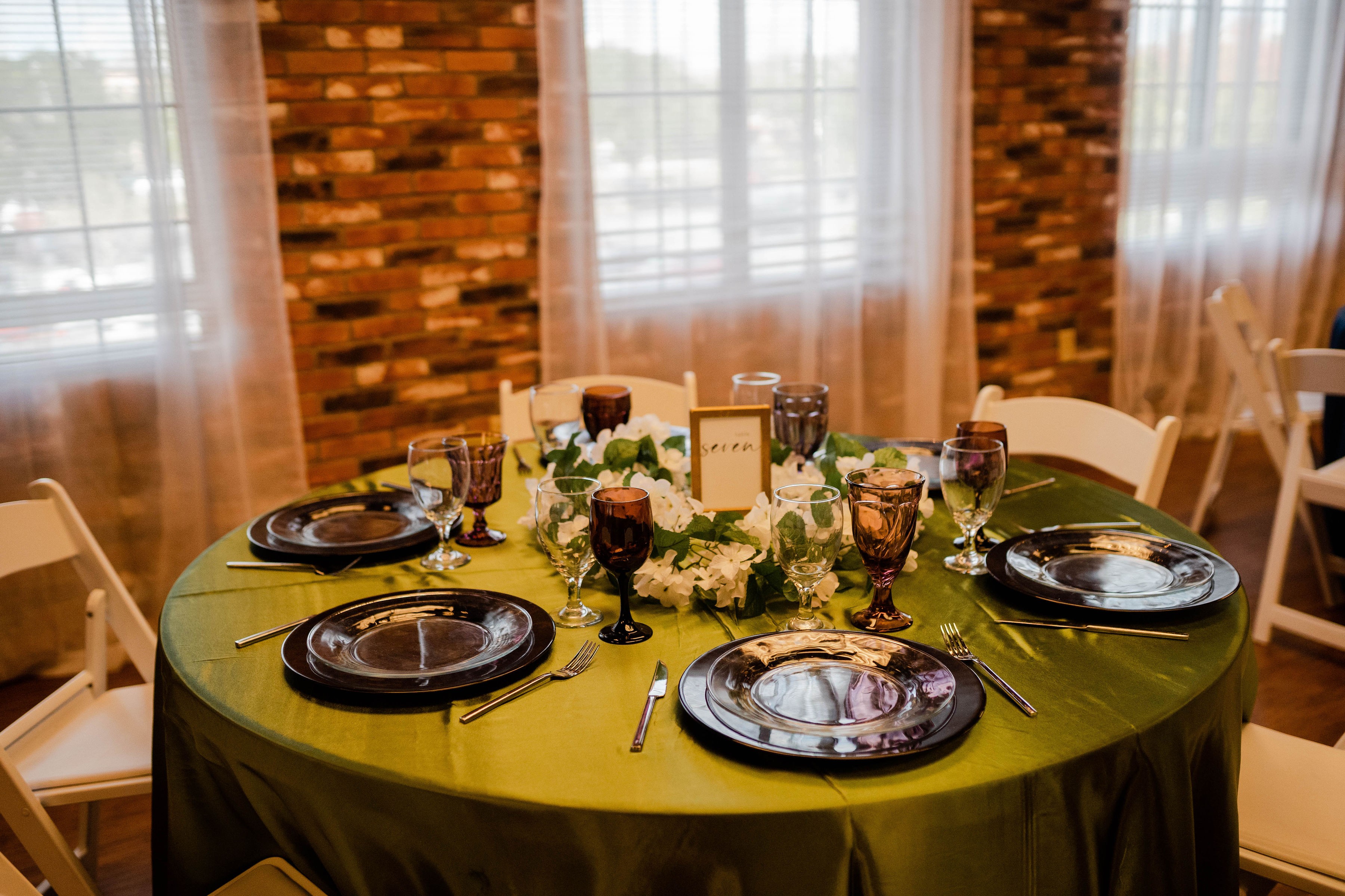 Round table with green tablecloth, set with black plates, glasses, and silverware, decorated with flowers and candles, in a room with brick walls and curtains.