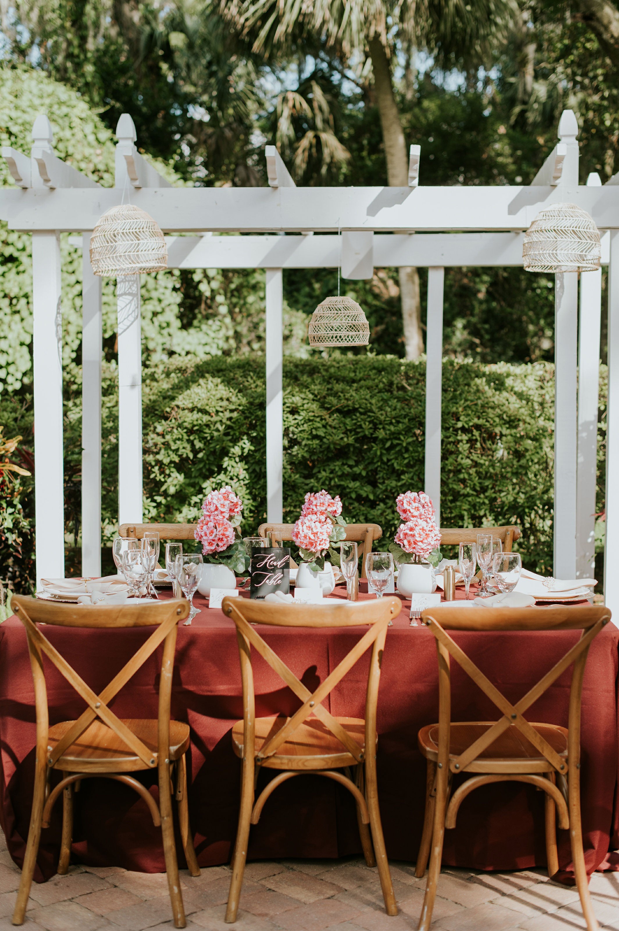 Outdoor table set with pink flowers, glassware, and chairs under a white gazebo, surrounded by greenery.