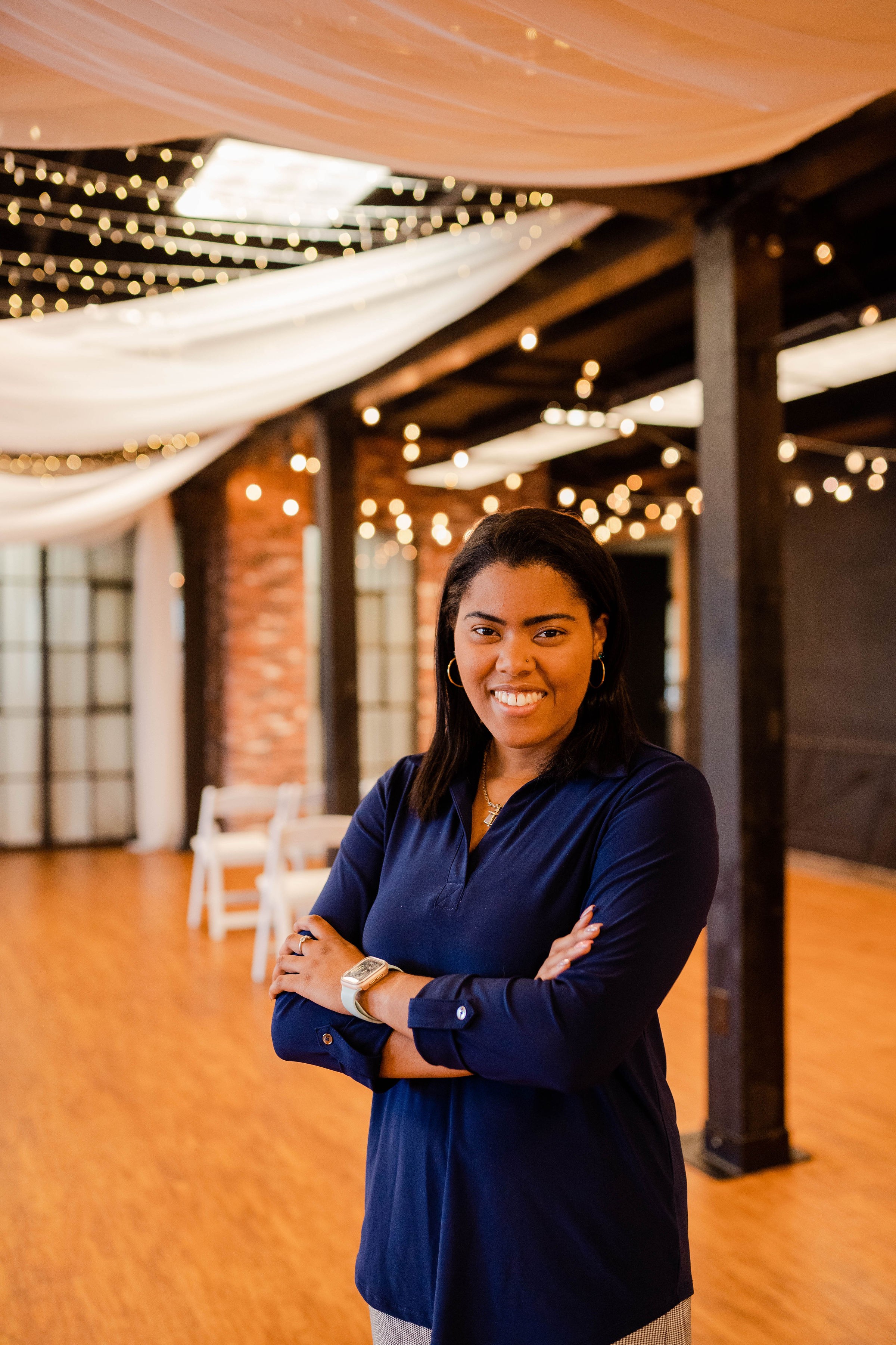 Smiling woman with crossed arms in a decorated event space with draped fabric and string lights.