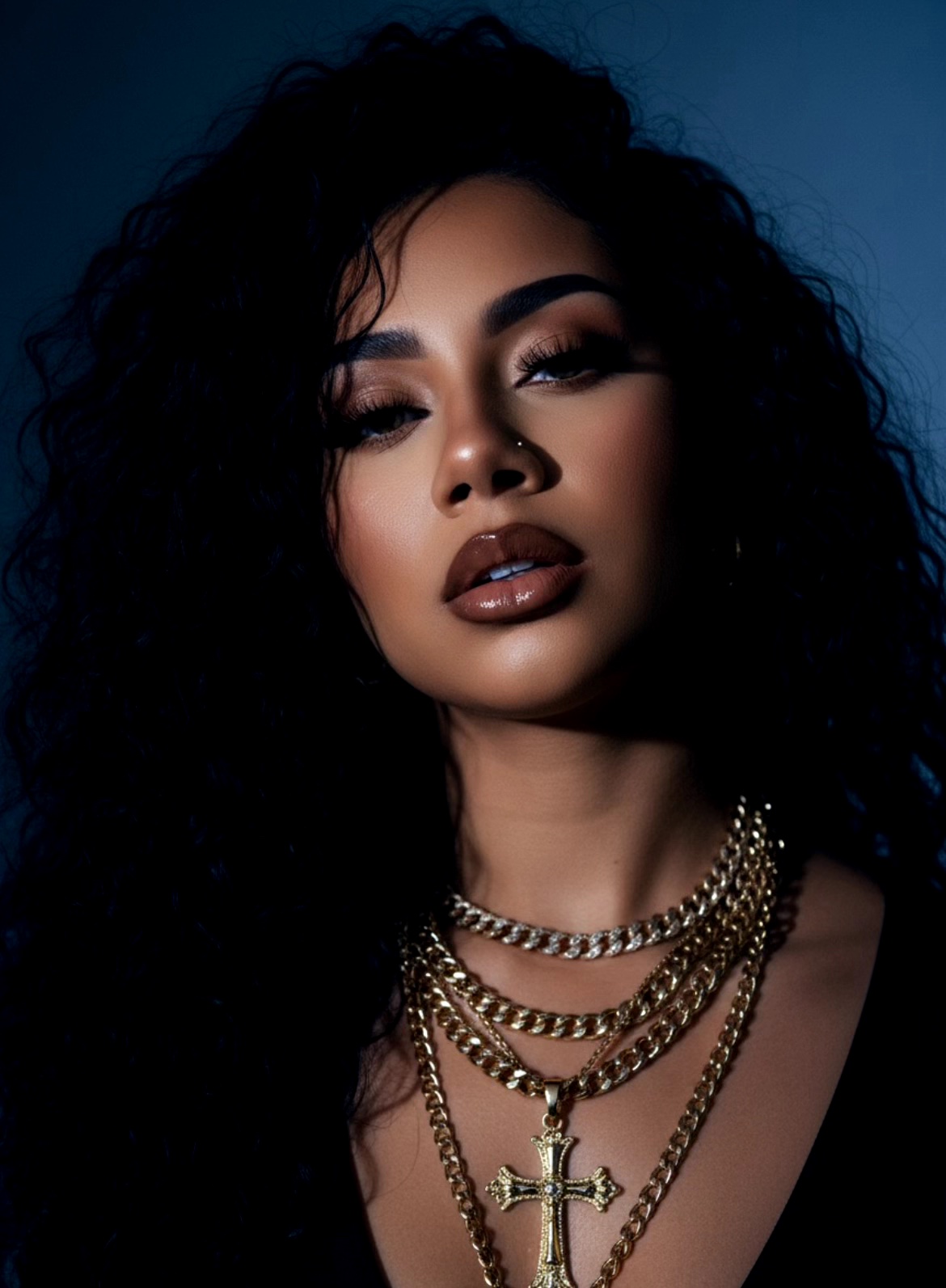 Portrait of a woman with curly hair, dark makeup, and layered gold necklaces against a dark background.