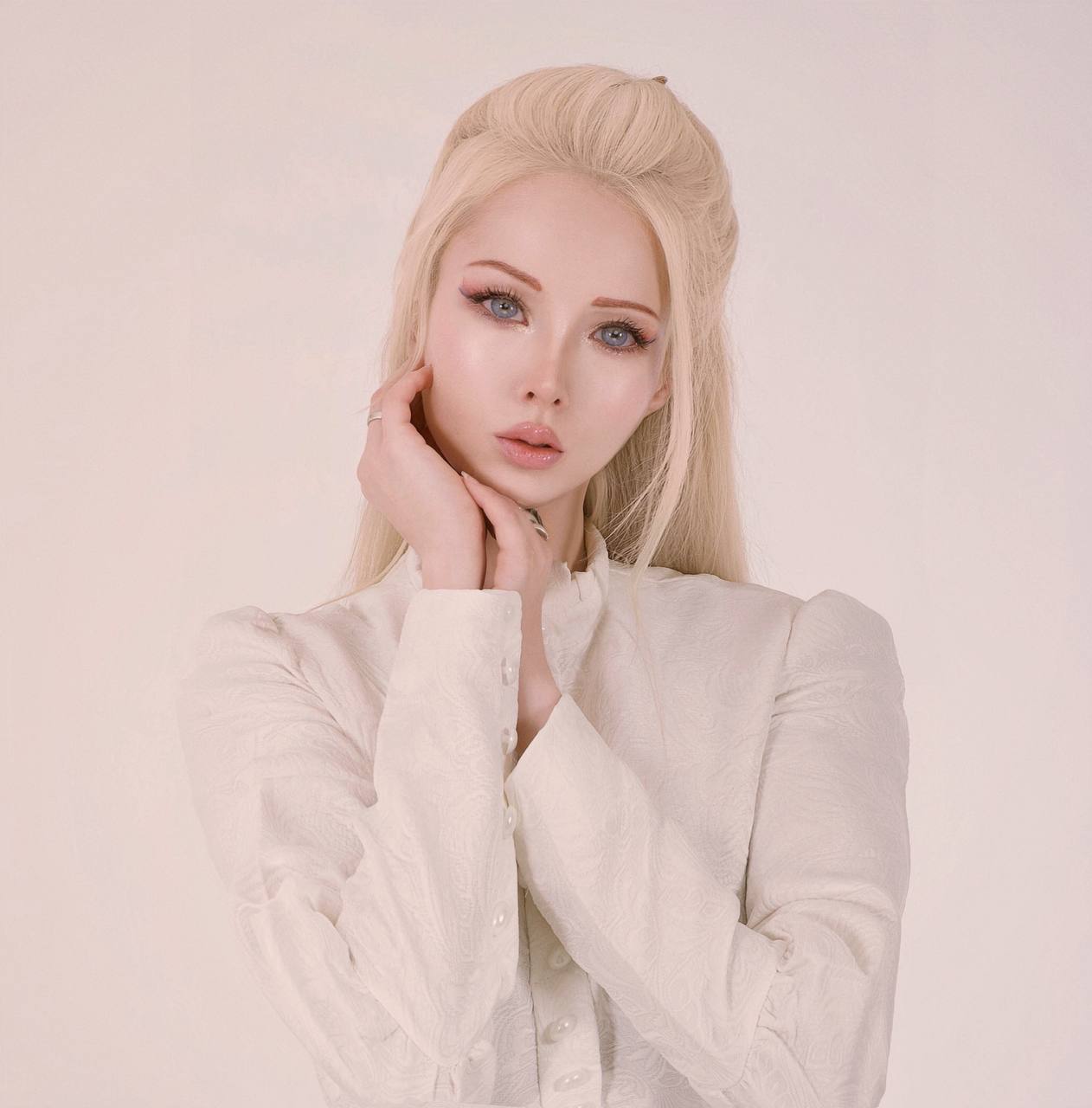 Young woman with platinum blonde hair and light makeup, wearing a light-colored top, posing with her hand near her face.