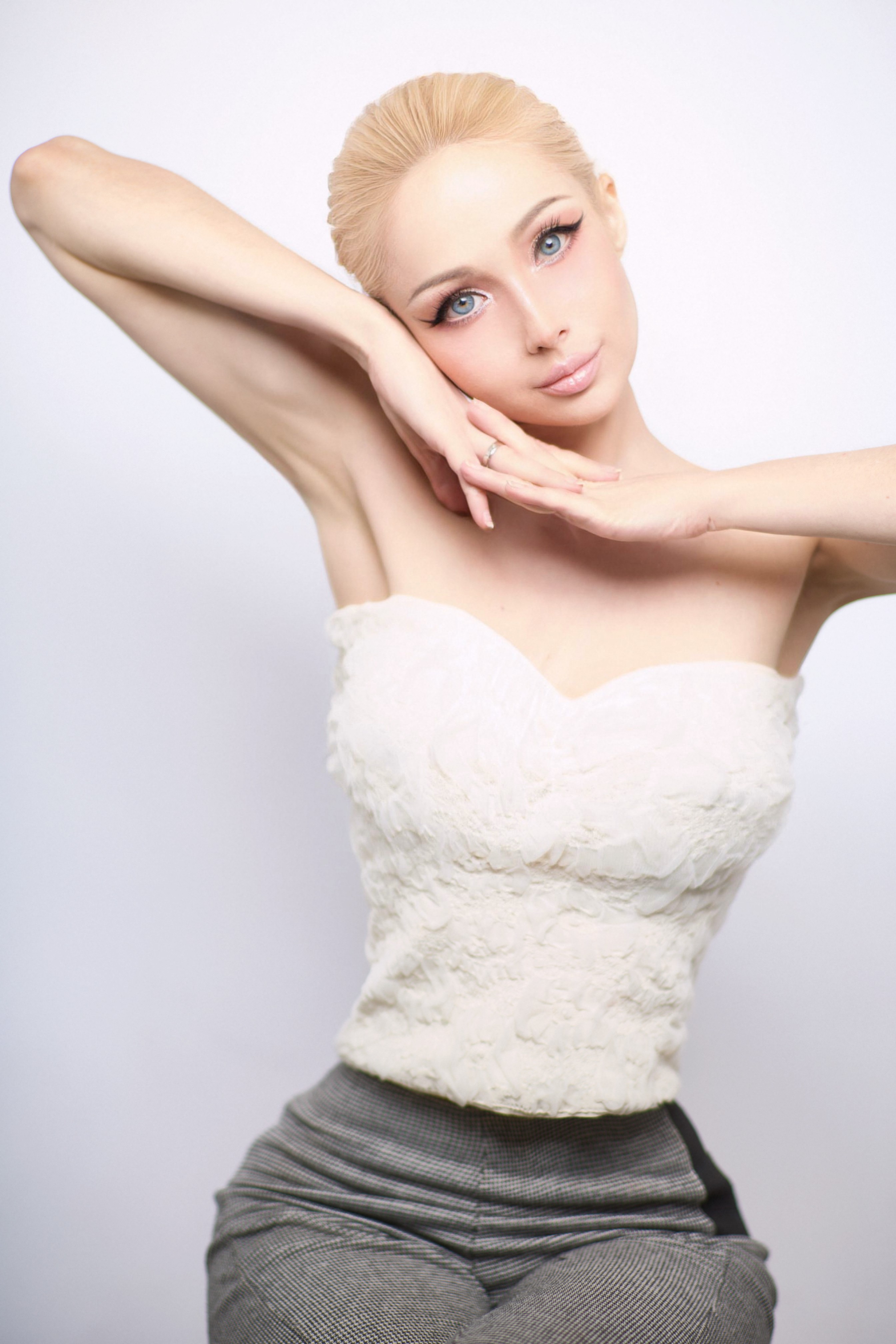Young woman with blonde hair and blue eyes poses with arms raised, wearing a white strapless top and gray pants.