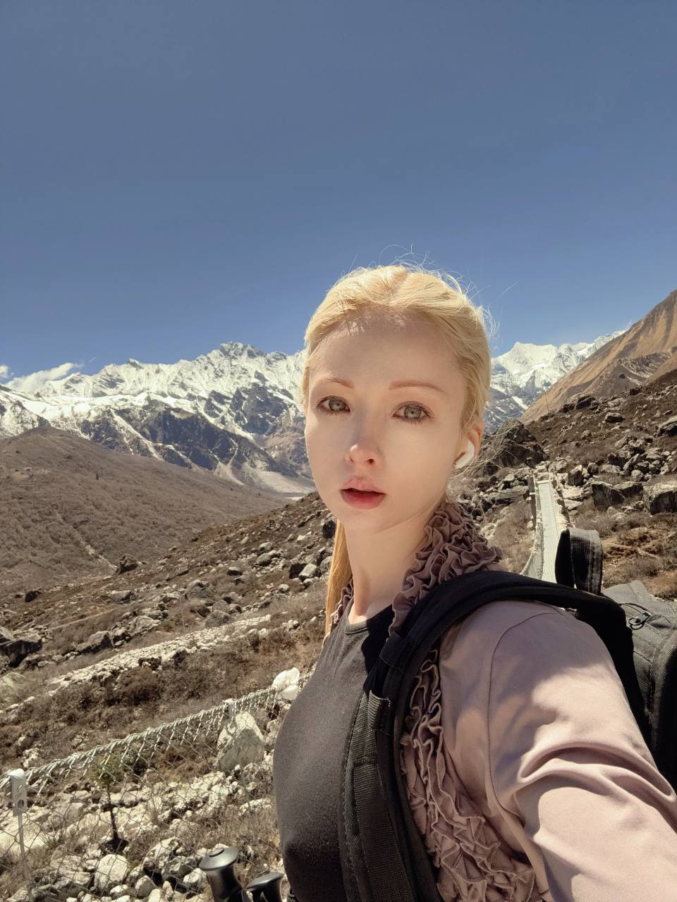 Young woman with blonde hair in a ponytail taking a selfie in mountainous terrain with snow-capped peaks and a clear blue sky.