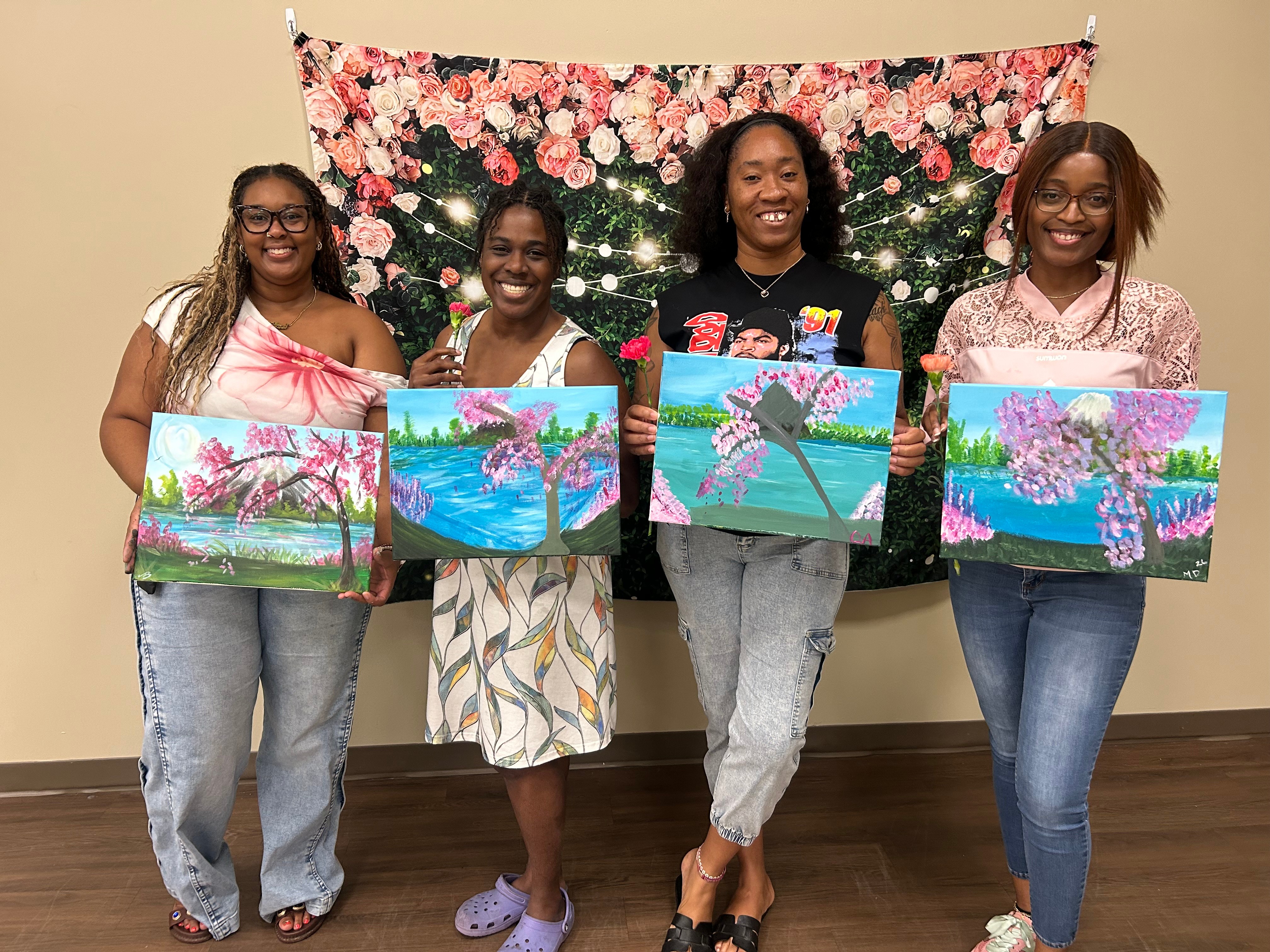 Four women stand indoors holding colorful paintings, with a floral backdrop behind them.
