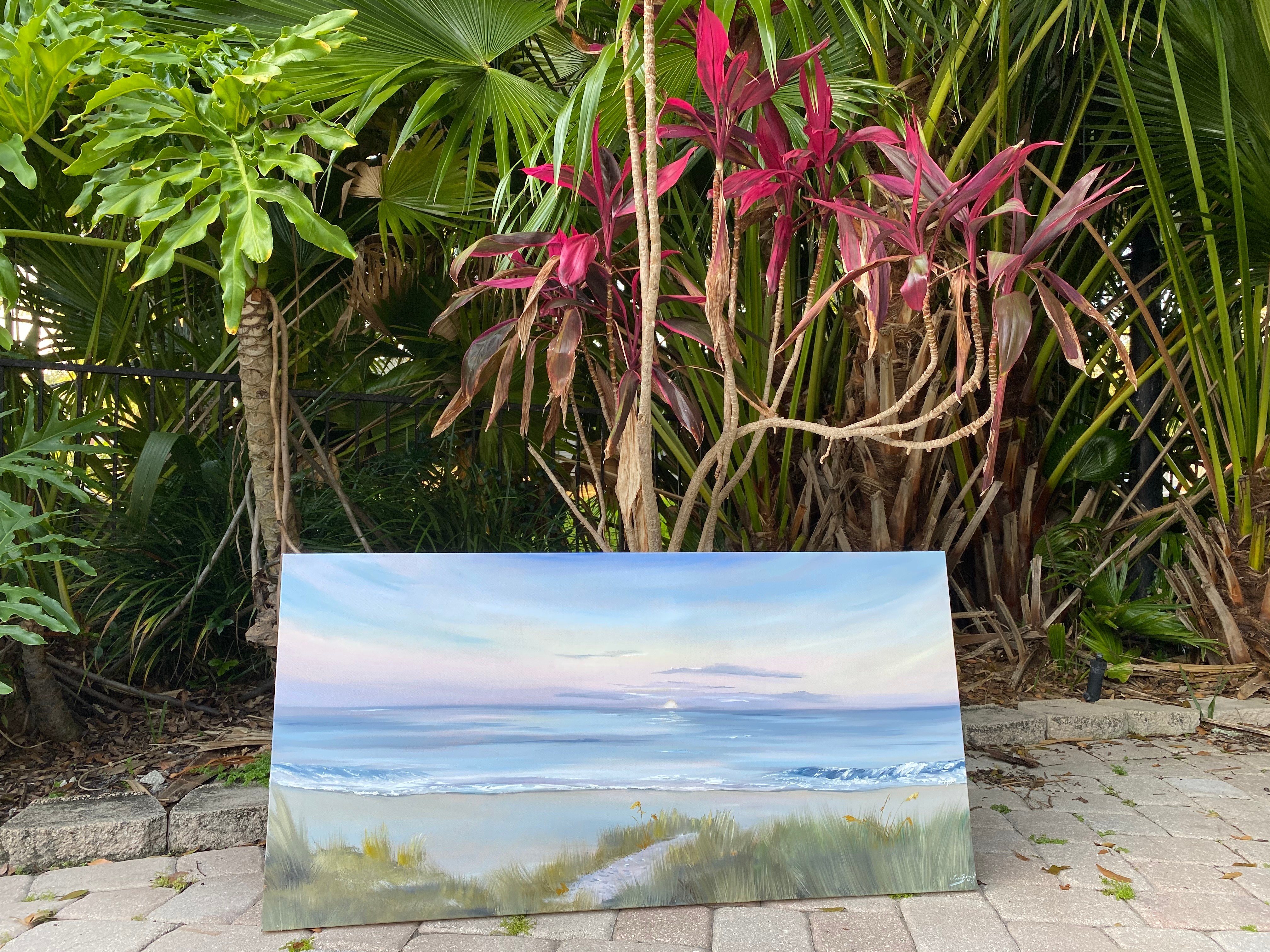 Landscape painting of a beach scene with sky, water, and greenery, placed on the ground among tropical plants.