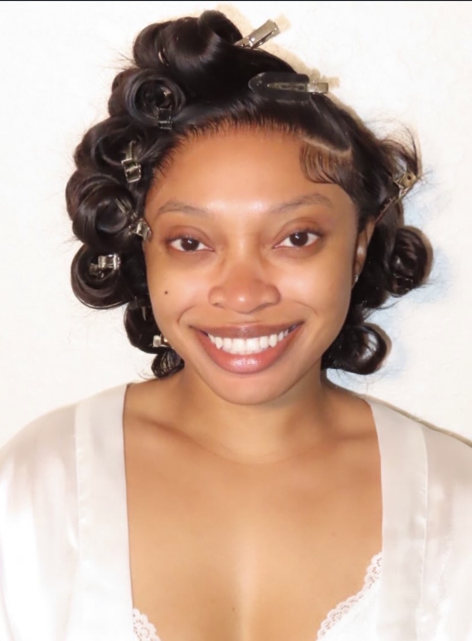 Young woman with styled hair, smiling, wearing a light-colored top, against a plain background.