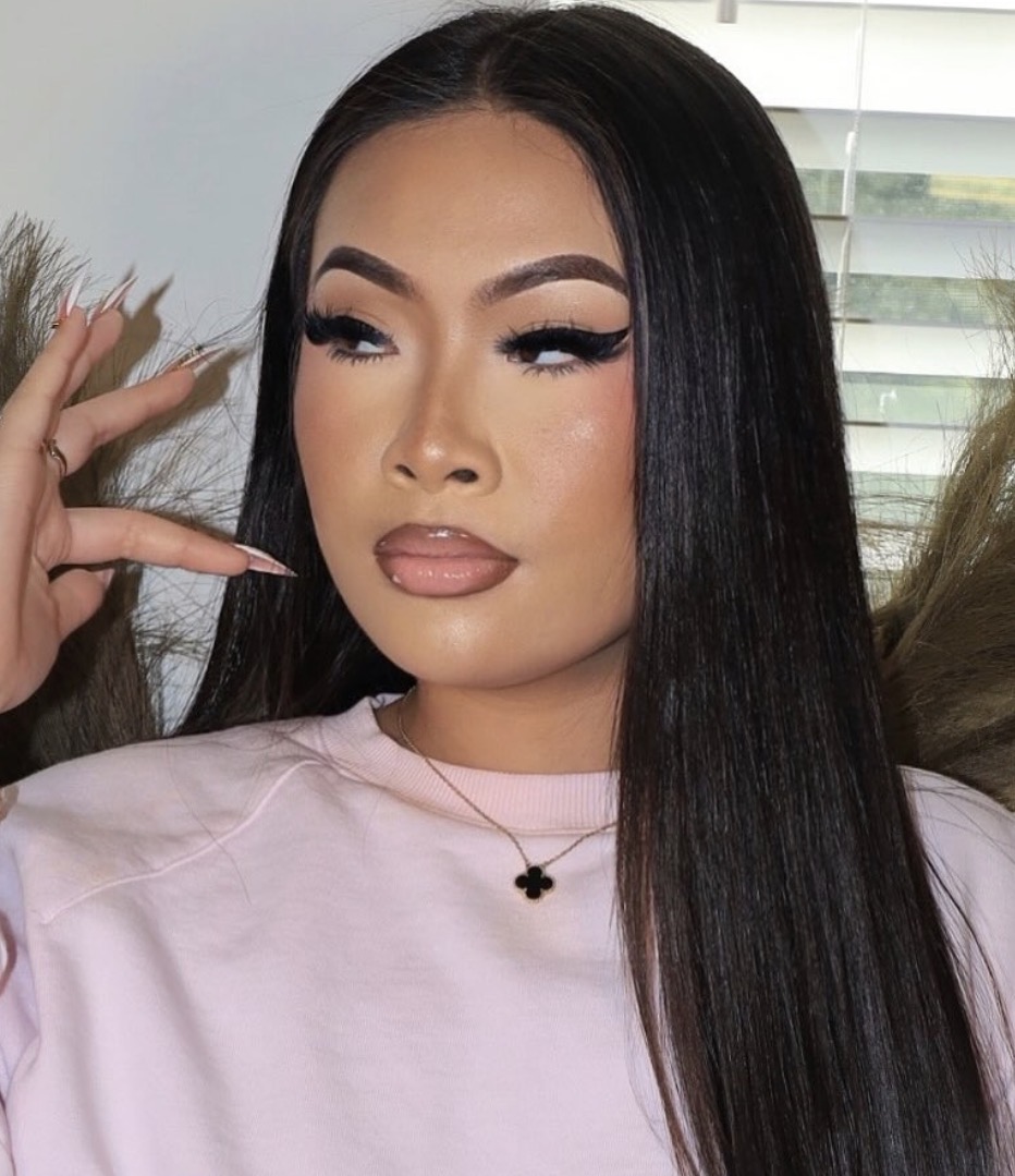 Woman with long black hair, makeup, and a pink shirt, posing indoors near a window with blinds.