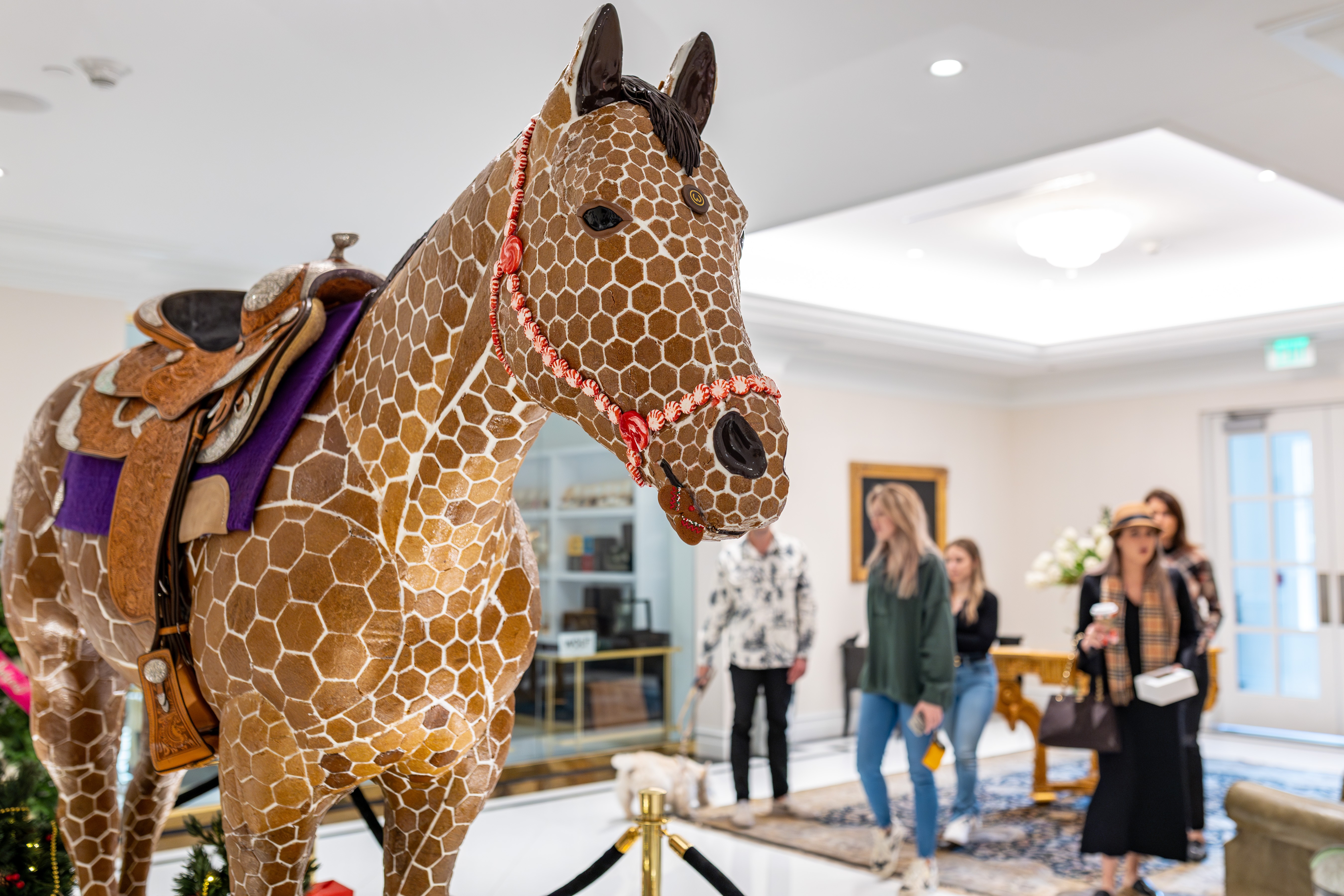 Giant giraffe-shaped sculpture with a saddle and harness in an indoor space with people in the background.