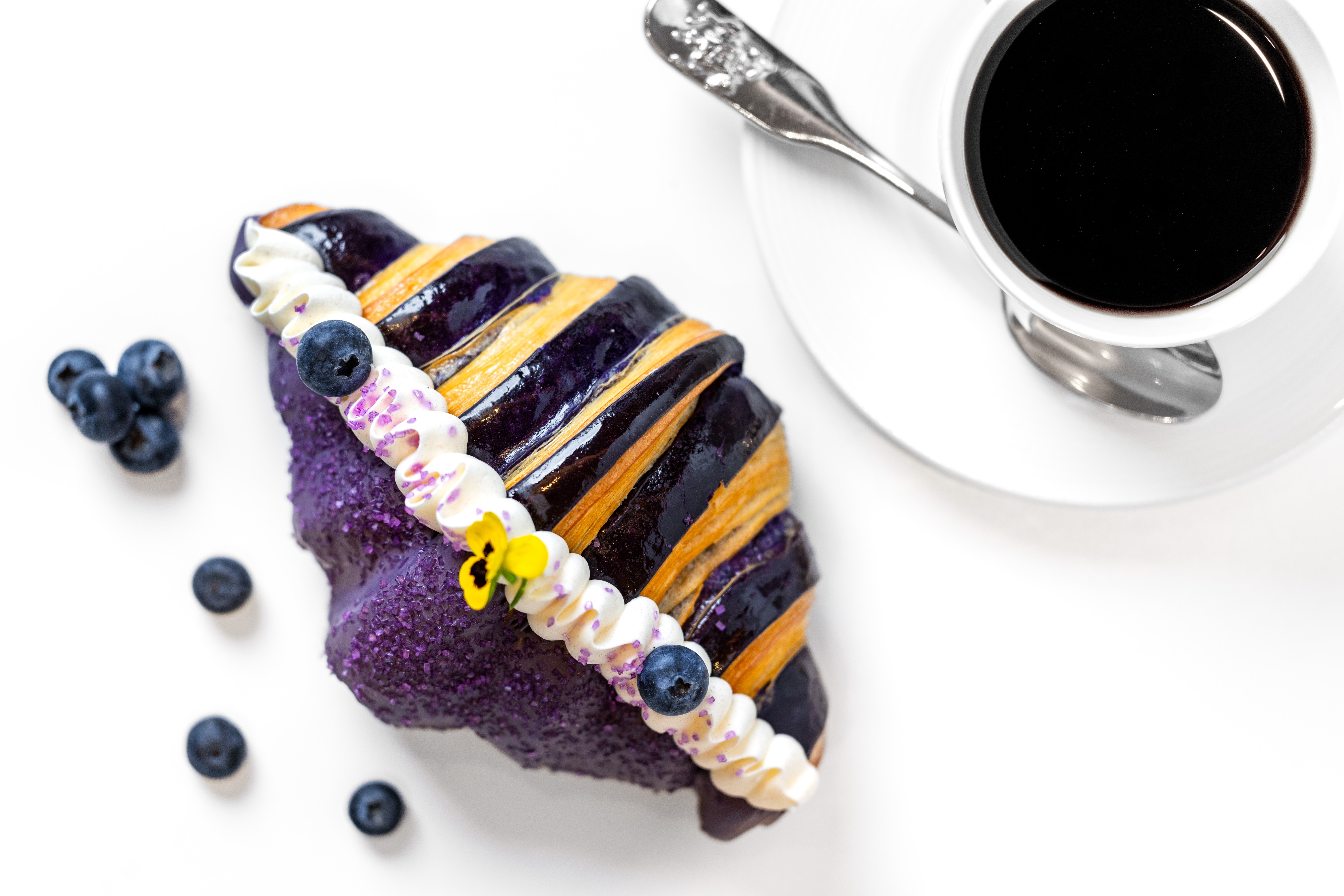 A slice of blueberry cake with purple icing, topped with blueberries and white cream, next to a cup of black coffee on a saucer with a spoon.