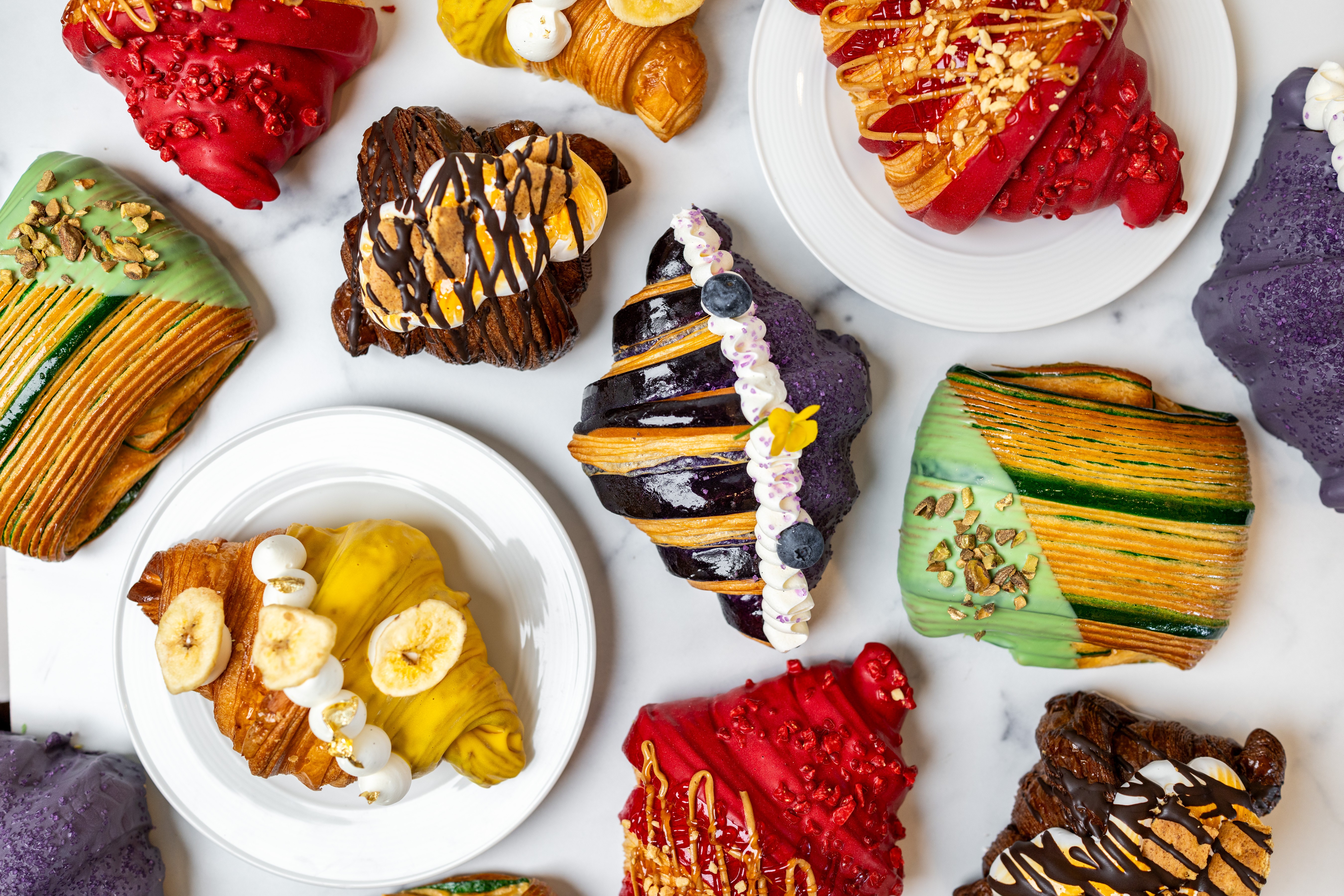 Assorted slices of colorful layered and decorated cakes and desserts on white plates, with some purple napkins nearby.