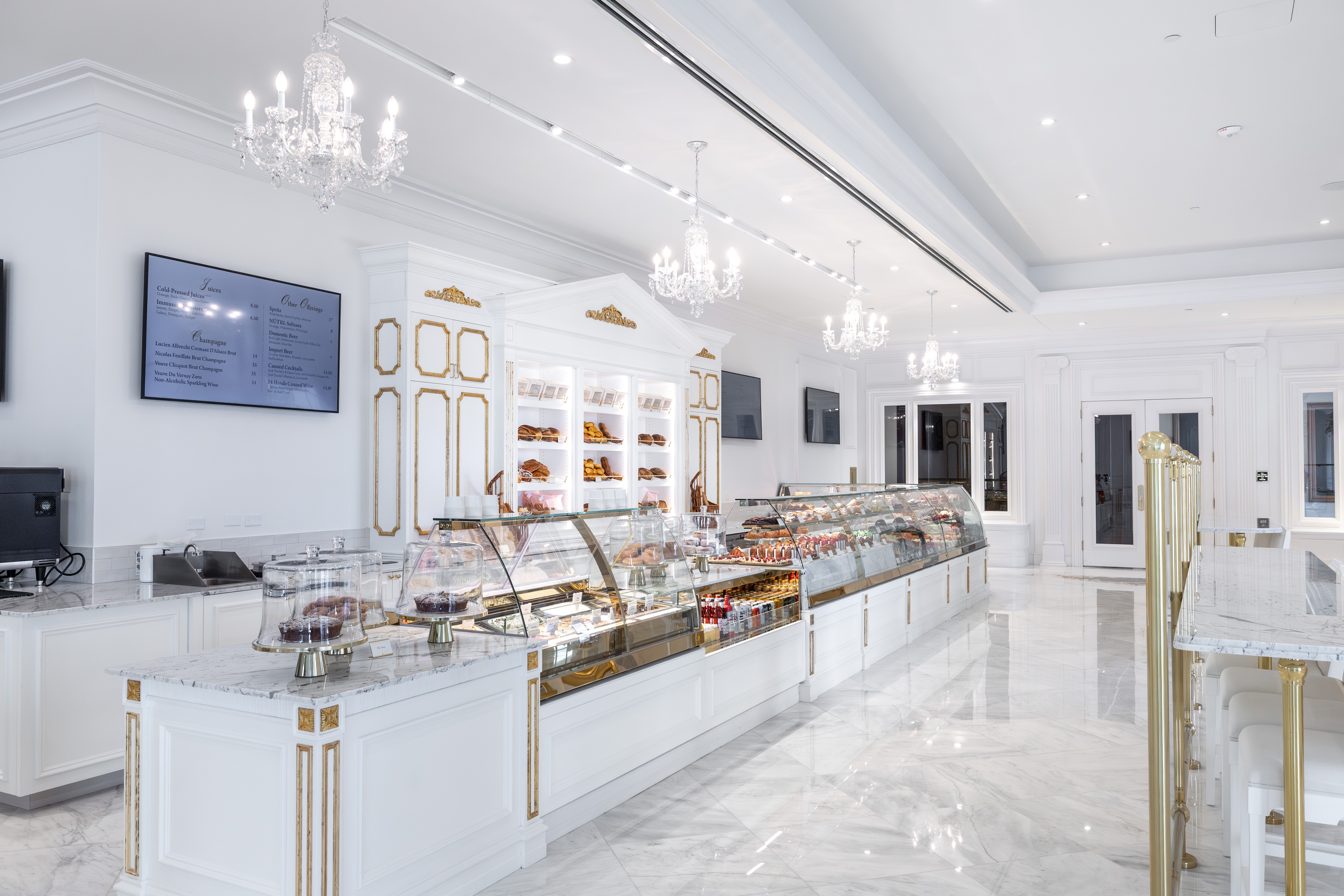 Elegant bakery interior with white walls, chandeliers, display case, and menu screens, bright and spacious with a luxurious design.