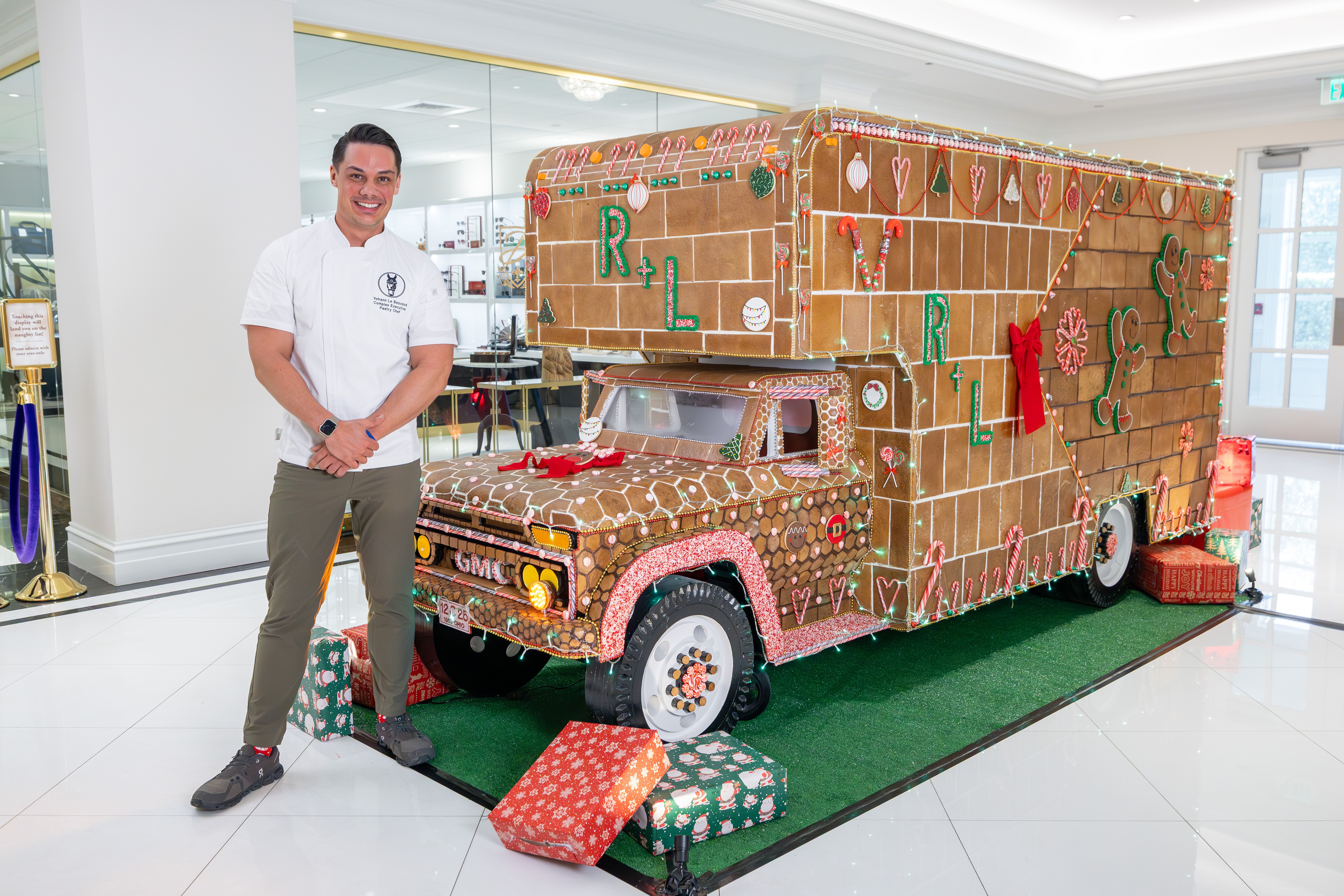 Person standing next to a large gingerbread house on a decorated vehicle, with presents nearby, in a bright indoor space.