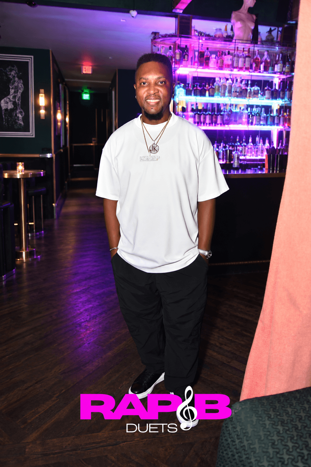 Young man standing in a bar with colorful illuminated shelves, wearing a white t-shirt and black pants, smiling.