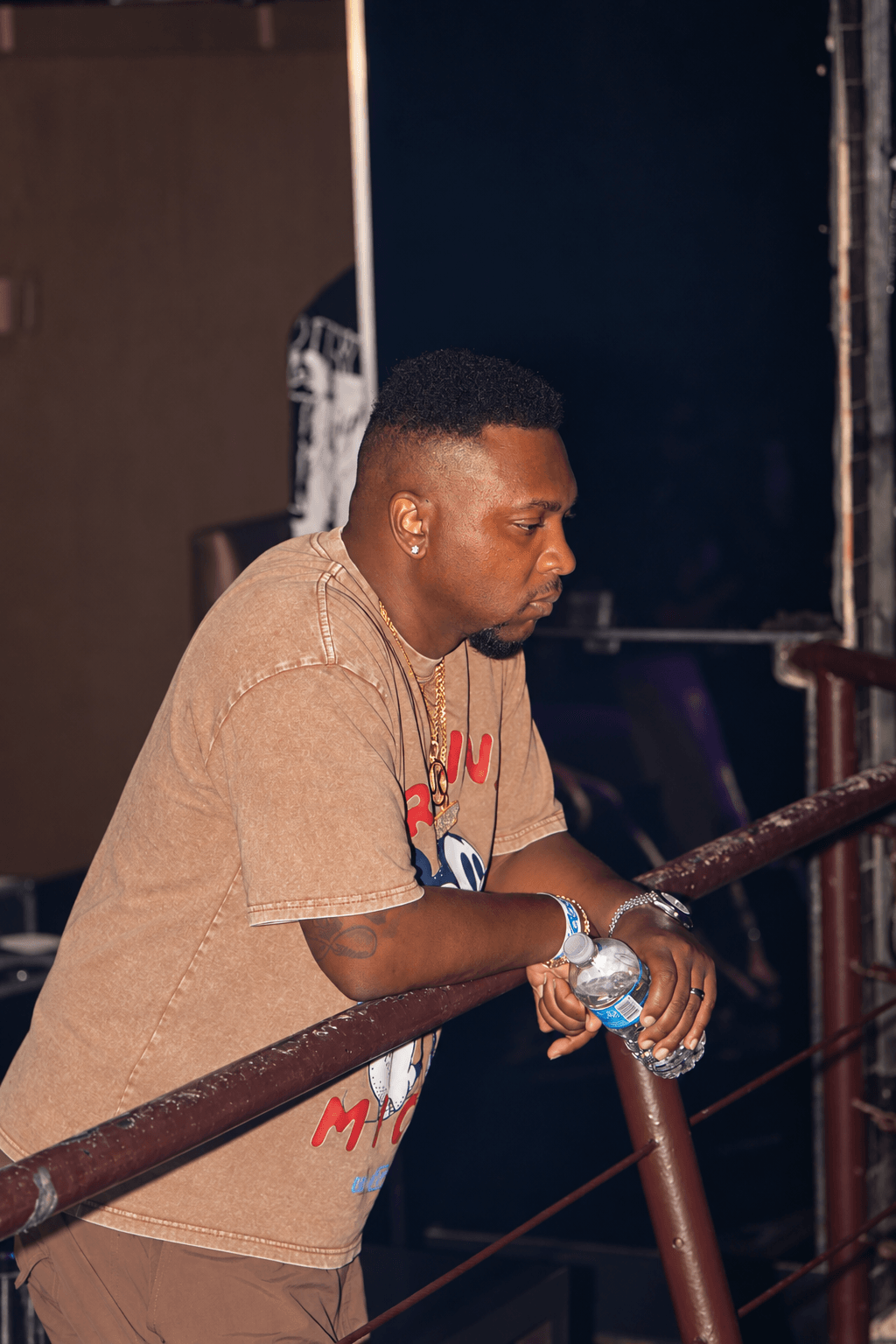 Young man with short hair wearing a beige T-shirt, holding a water bottle, leaning on a railing indoors.