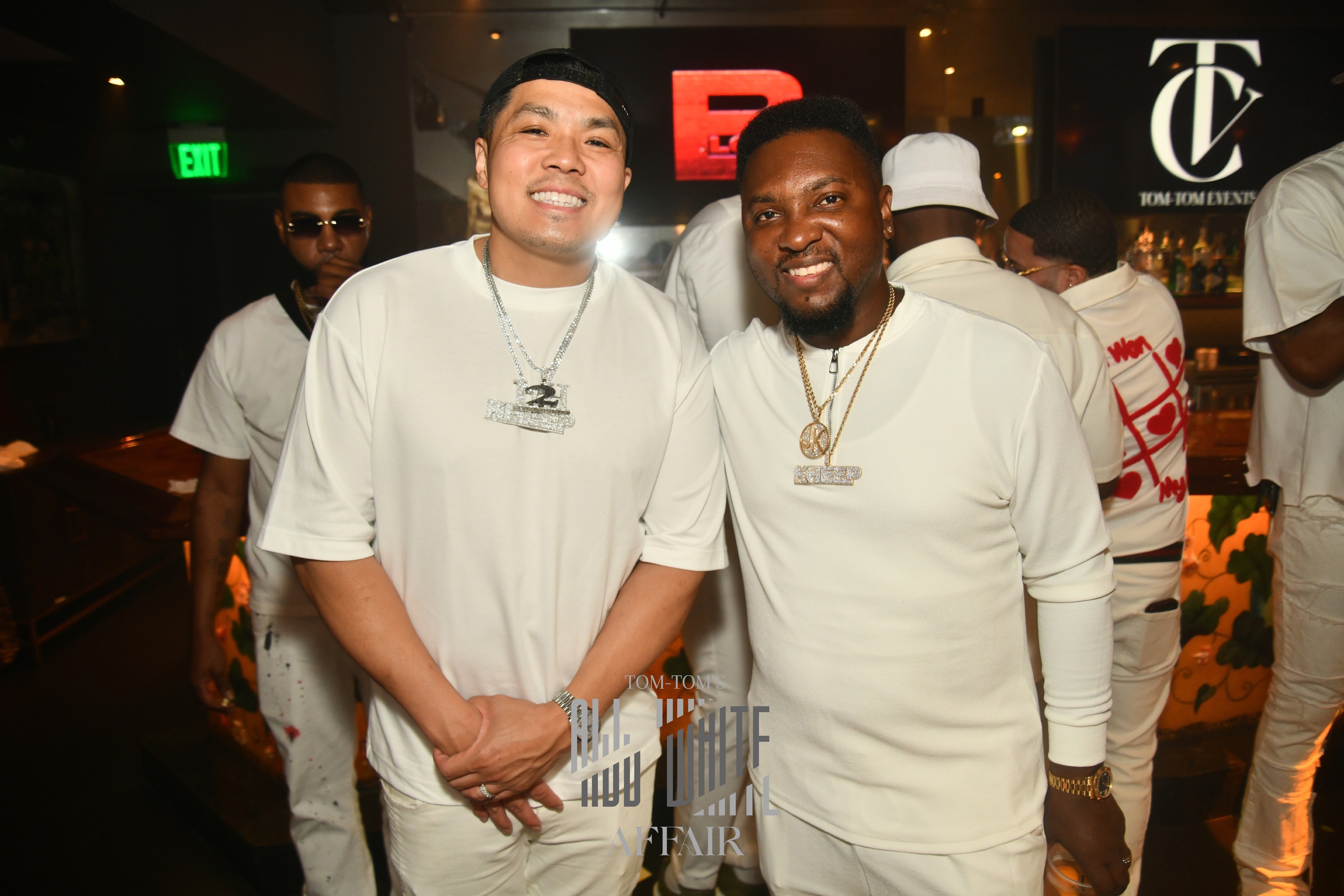 Two smiling men in white shirts pose together at an indoor event, with other people and logos in the background.