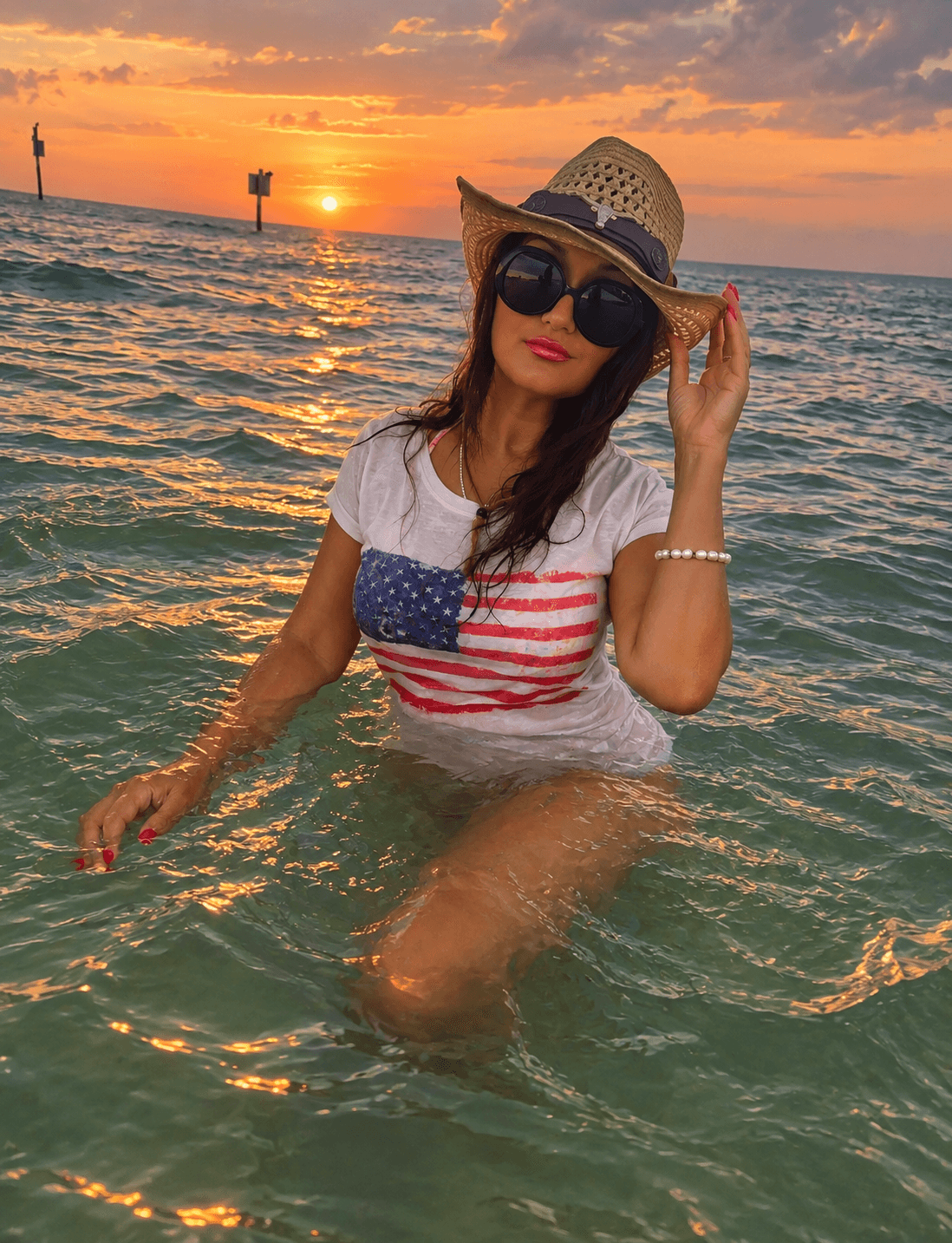 Woman in water wearing sunglasses, straw hat, and American flag shirt at sunset, with ocean and sky in background.