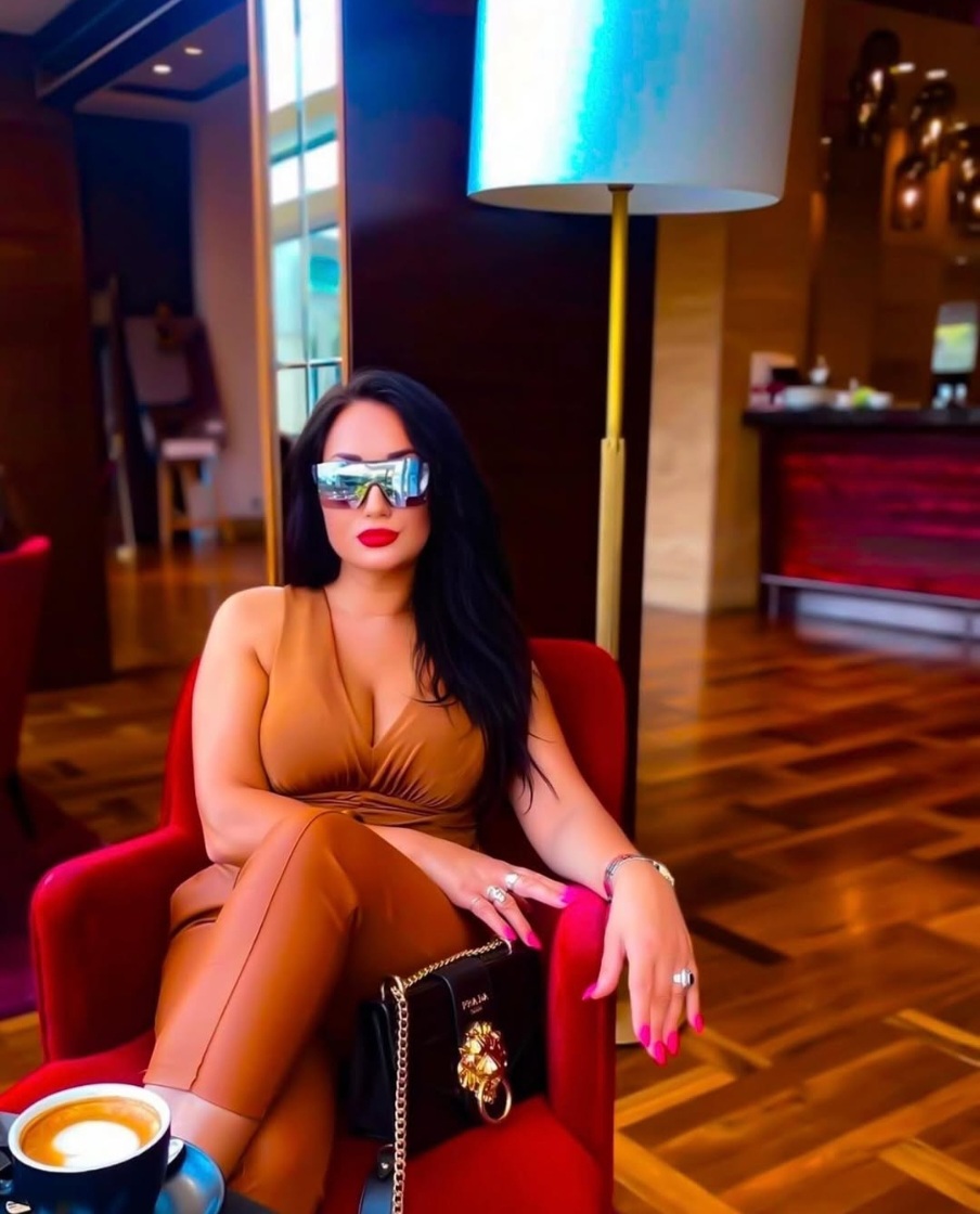 Woman with long dark hair and red lipstick sitting on a red chair in a room with wooden floors and a tall lamp.