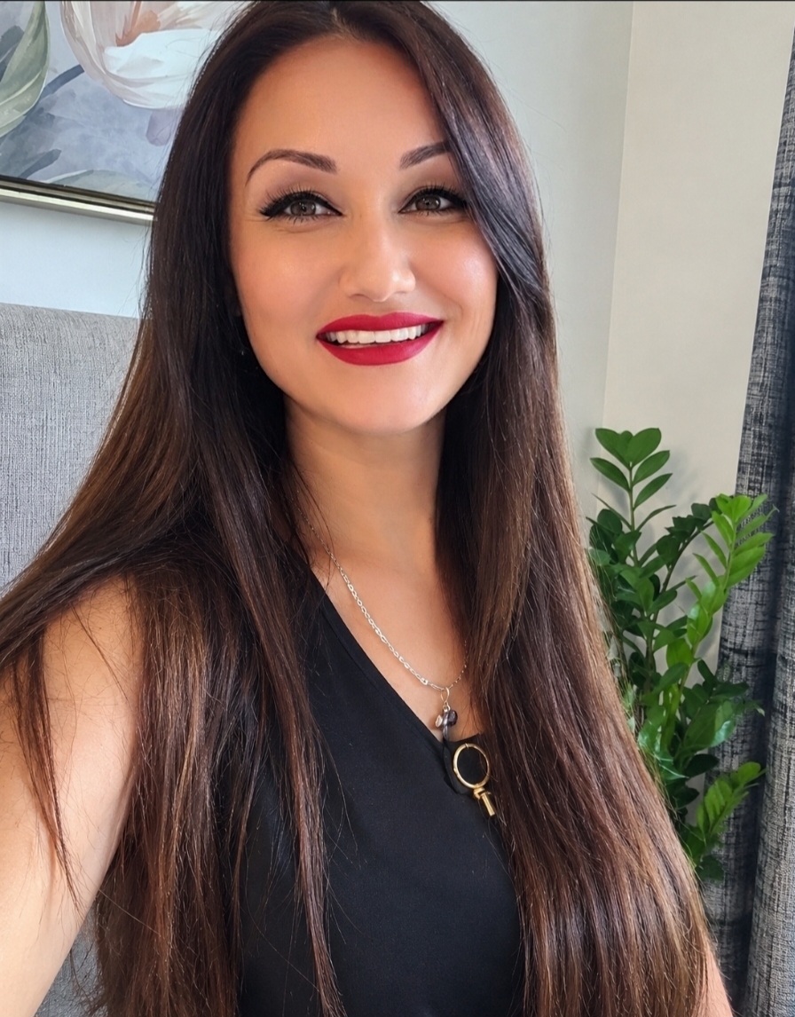 Young woman with long brown hair, red lipstick, smiling, wearing a black top and necklace, indoors with a plant in the background.