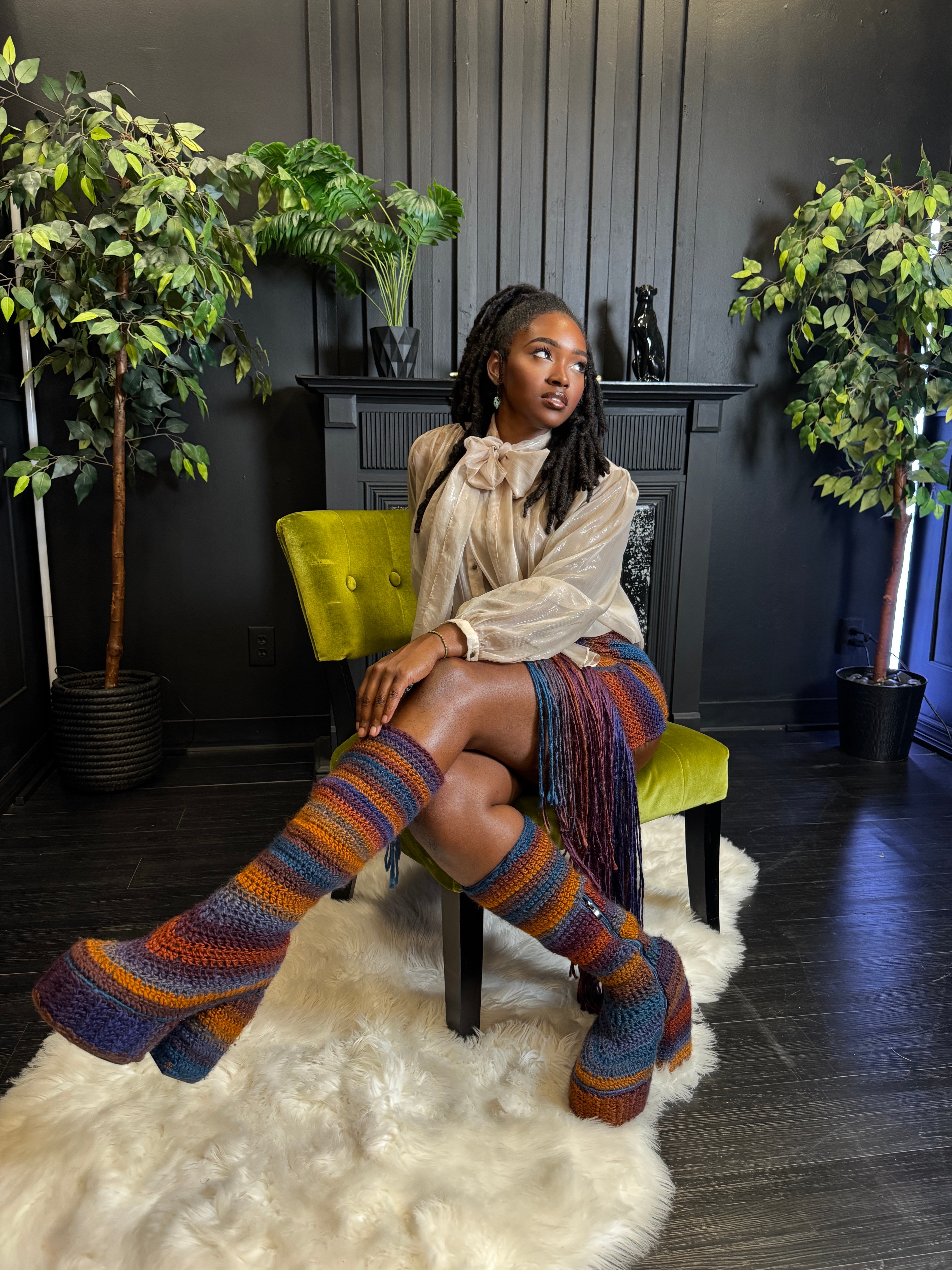 Young woman with long hair sitting on a yellow chair, wearing a beige blouse and colorful striped leg warmers, in a room with plants and dark walls.