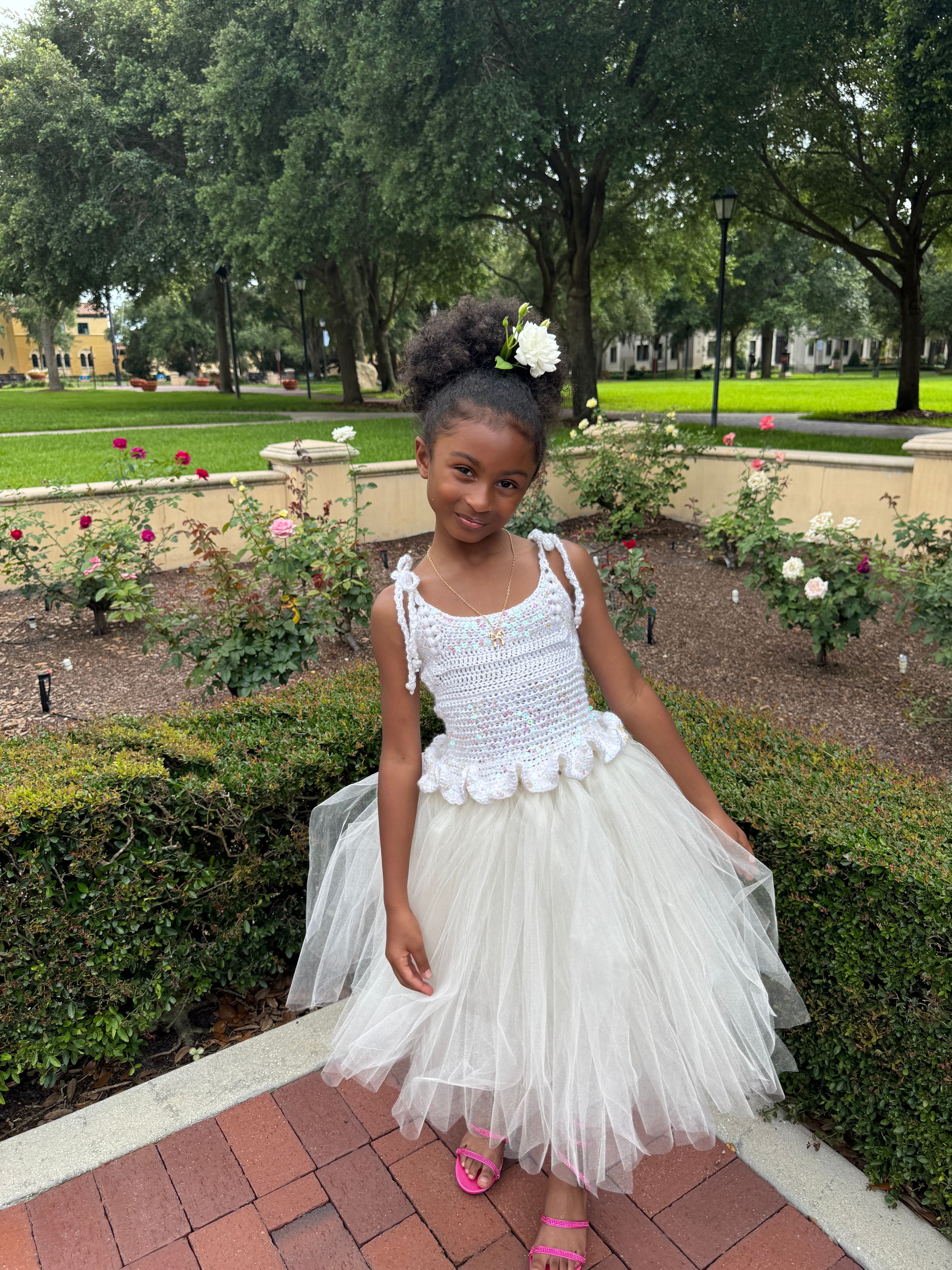 Young girl in a white dress with a tulle skirt, standing outdoors on a brick path, surrounded by greenery and flowers.