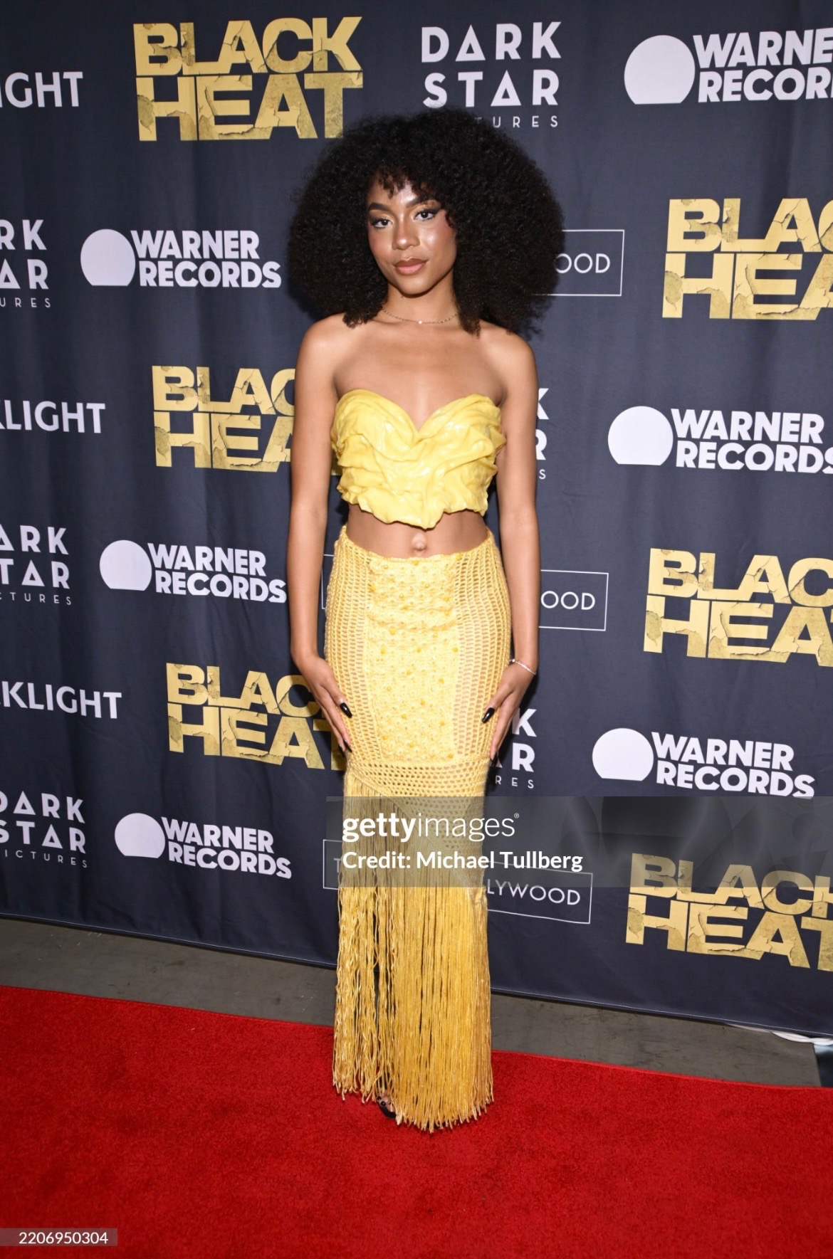 Woman with curly hair in a yellow two-piece outfit with fringe skirt, standing on red carpet at event, background with logos.