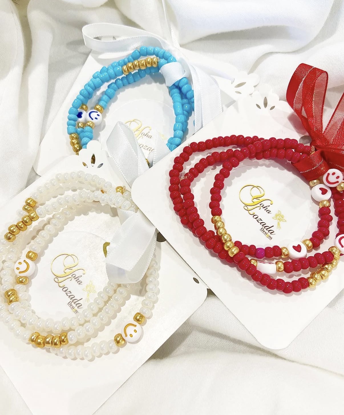 Three beaded bracelets with colorful beads and charms, displayed on white cards with gold lettering, on a white fabric background.