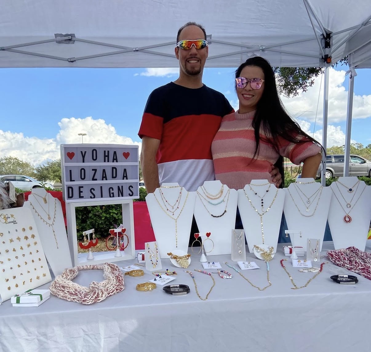Two people stand behind a jewelry display table under a canopy, with a sign reading 'Y O H A ♥ LOZADA DESIGNS' in the background.