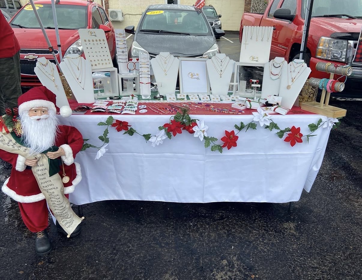 Table with holiday decorations and gift items, decorated with red poinsettias and garland, outdoors with parked cars in background.