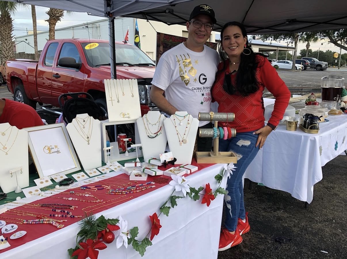 Two people stand behind a jewelry display at an outdoor market, with tables of jewelry and decorations in the background.