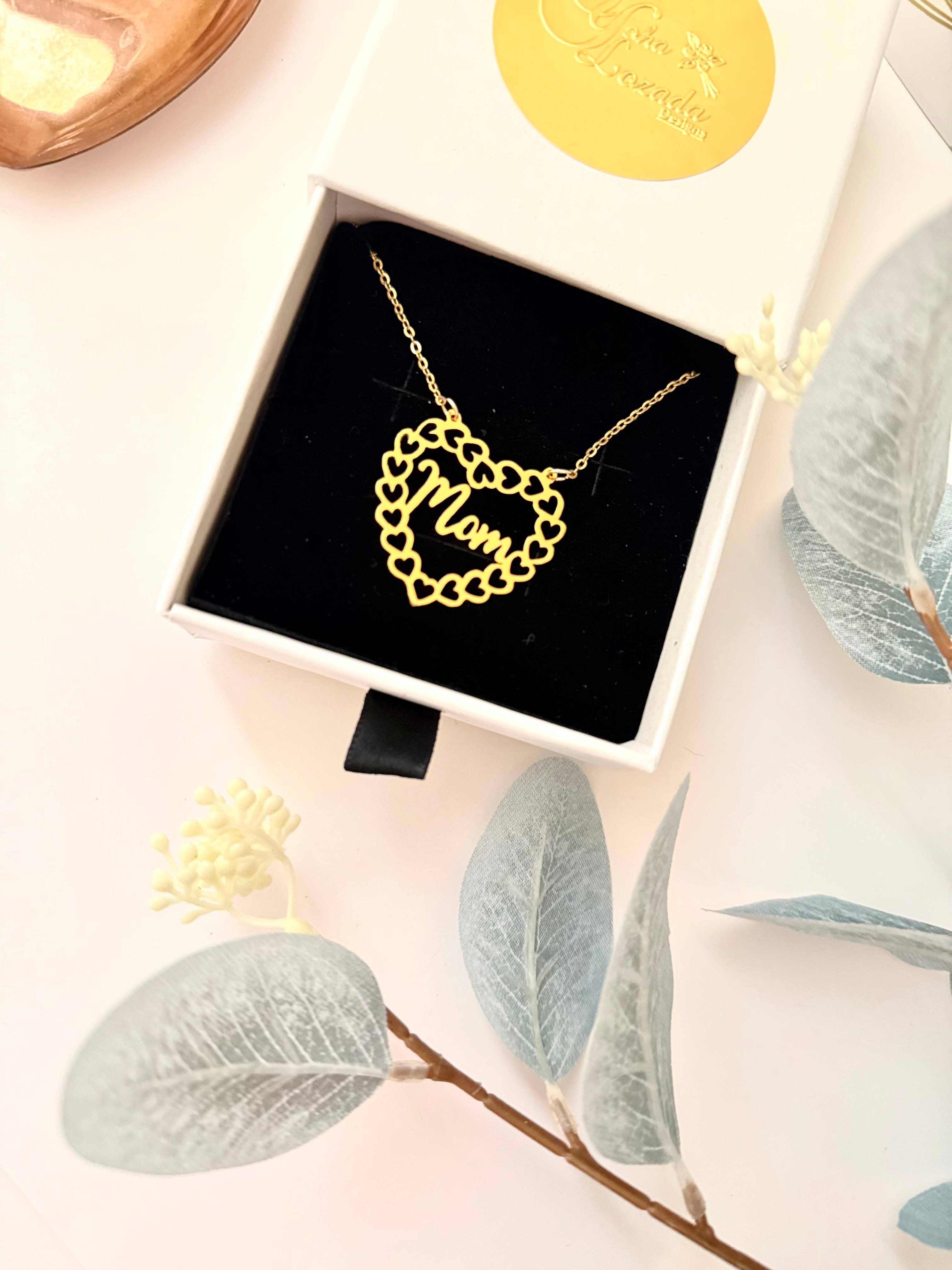 Gold necklace with a heart-shaped pendant inside a white box, surrounded by gray leaves and a yellow flower.