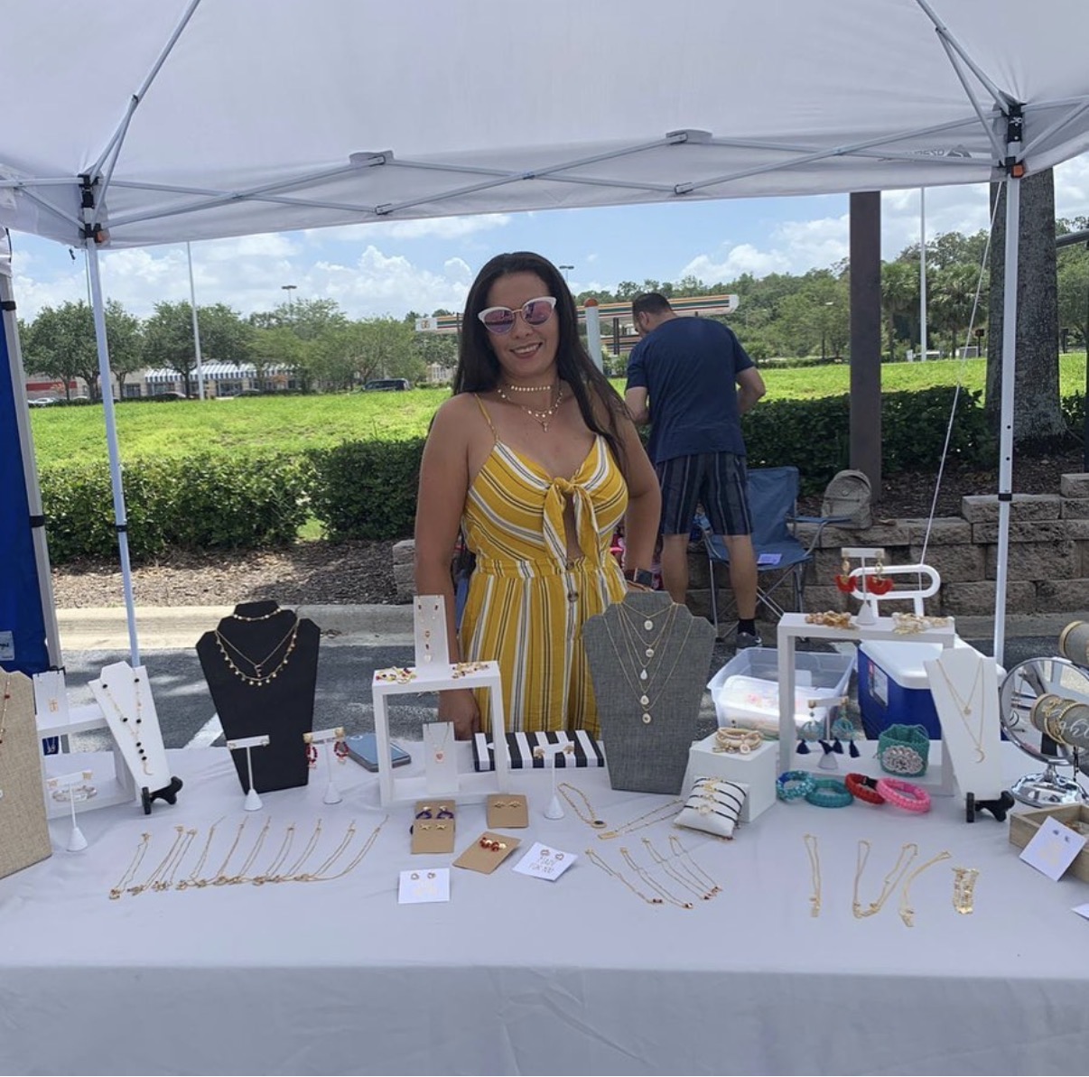 Woman in yellow striped dress stands behind jewelry display table outdoors, with people and greenery in background.