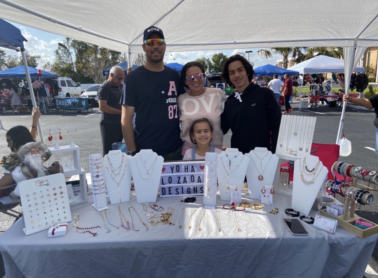Four people stand behind a jewelry display table at an outdoor market, with tents and shoppers in the background.
