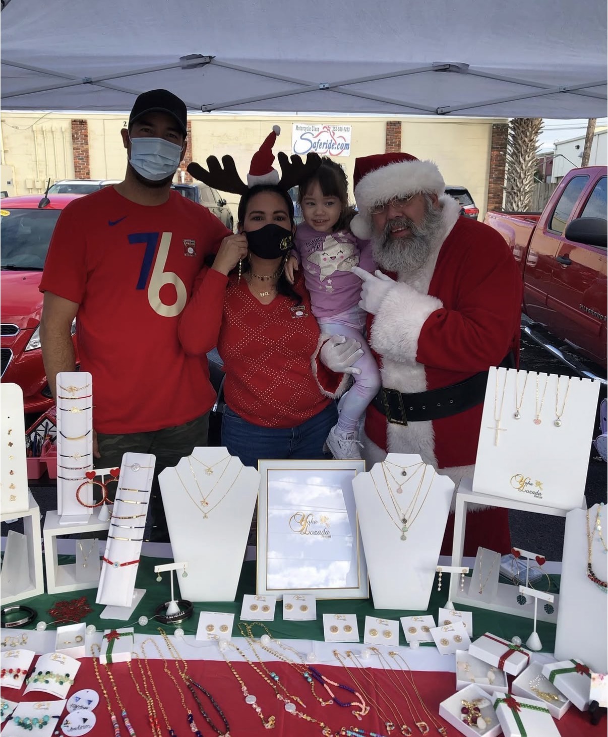 Four people, including a person dressed as Santa Claus, stand behind a table with jewelry displays at an outdoor event.