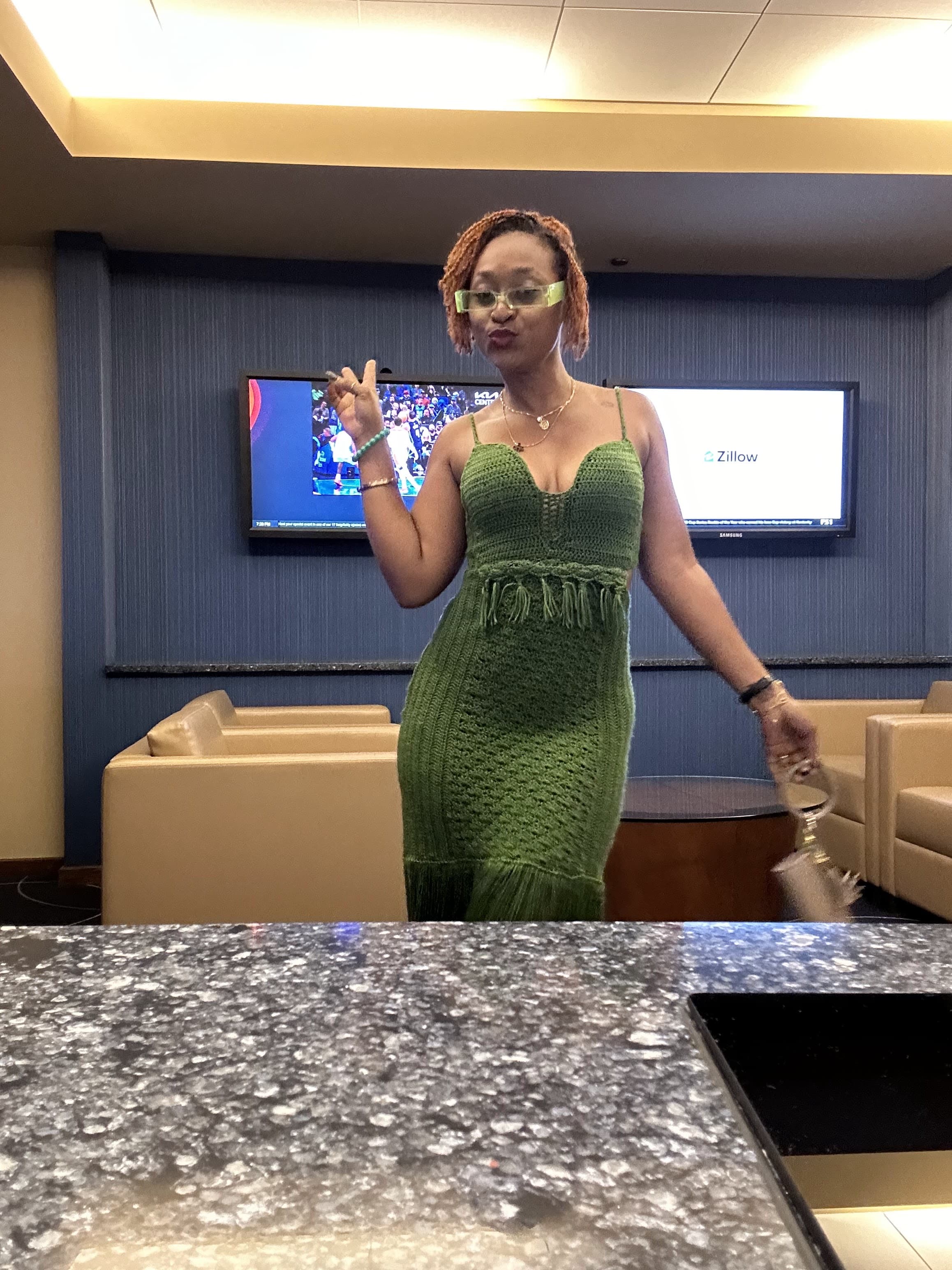 Woman in green dress standing in a room with TV screens, holding a drink, with beige furniture and a granite countertop.