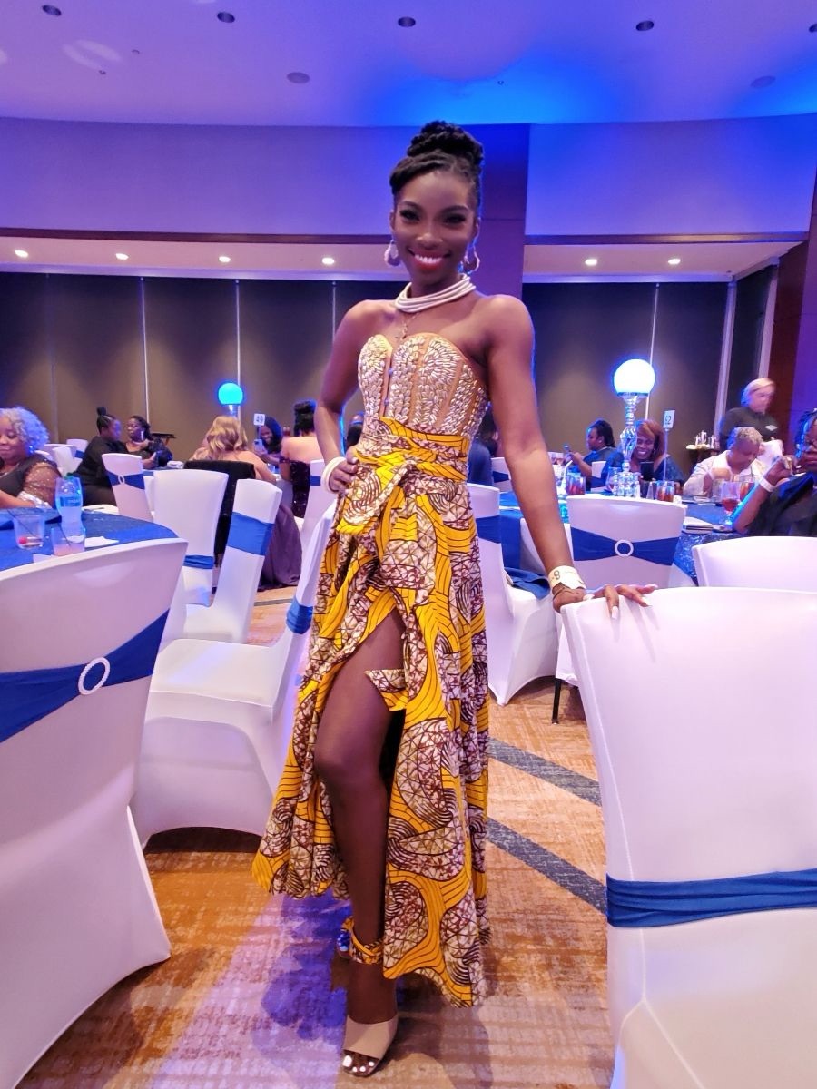 Woman in colorful dress standing in a decorated banquet hall, smiling, with tables and guests in background.