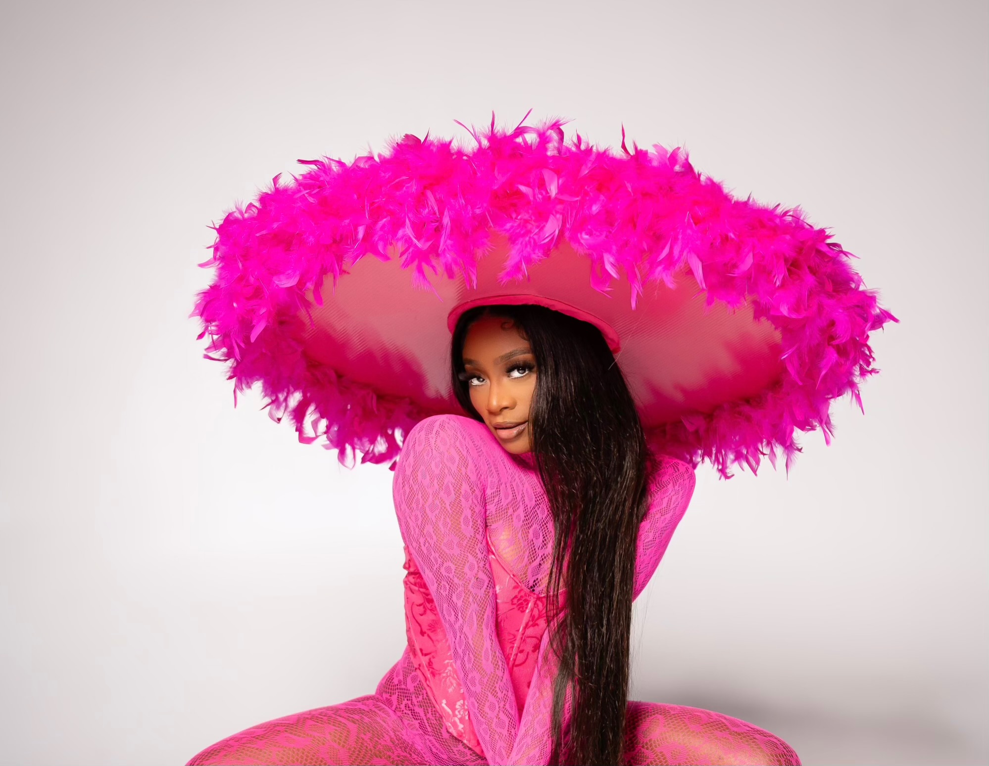 Woman in pink outfit with large pink feathered hat, sitting against a plain background.