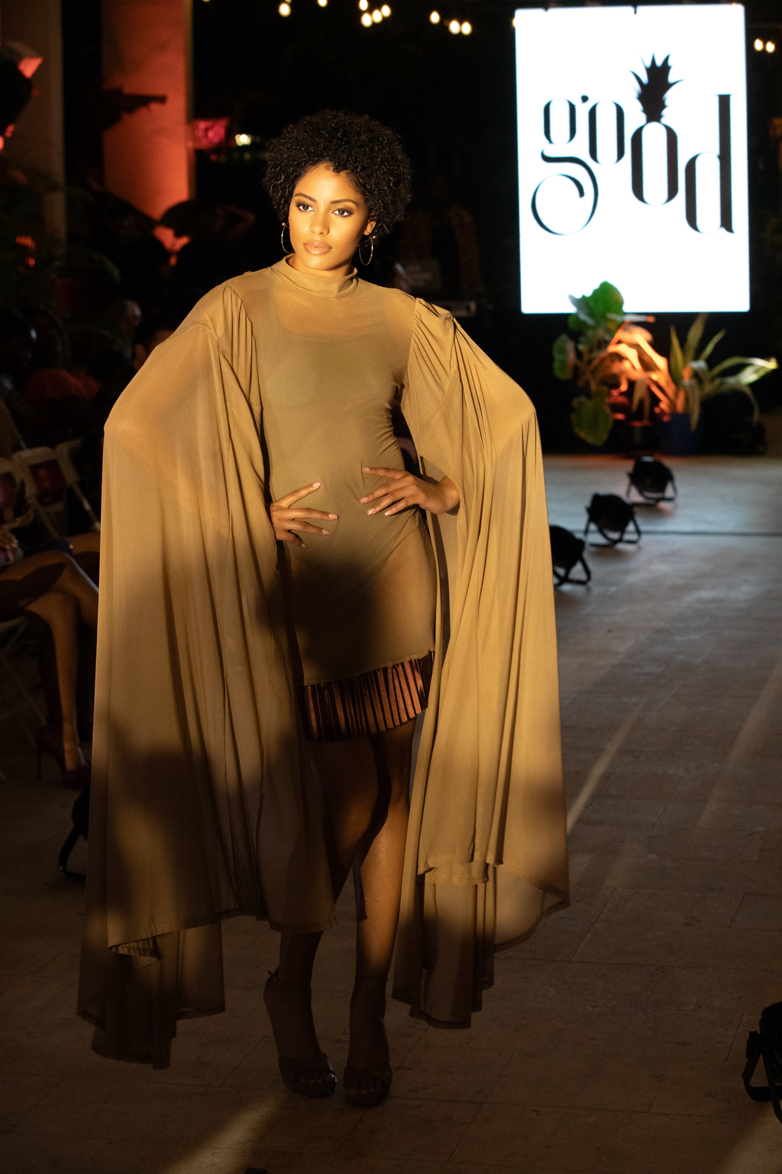 Model walking on runway in beige outfit with flowing cape, audience in background, illuminated sign reading 'GOD' behind.