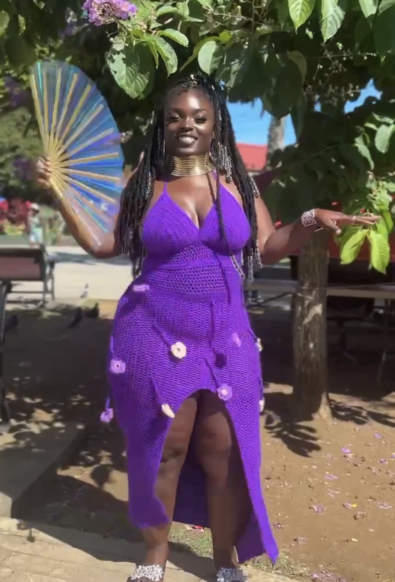Woman in purple dress holding a fan outdoors, standing near trees and benches, smiling, sunny day.