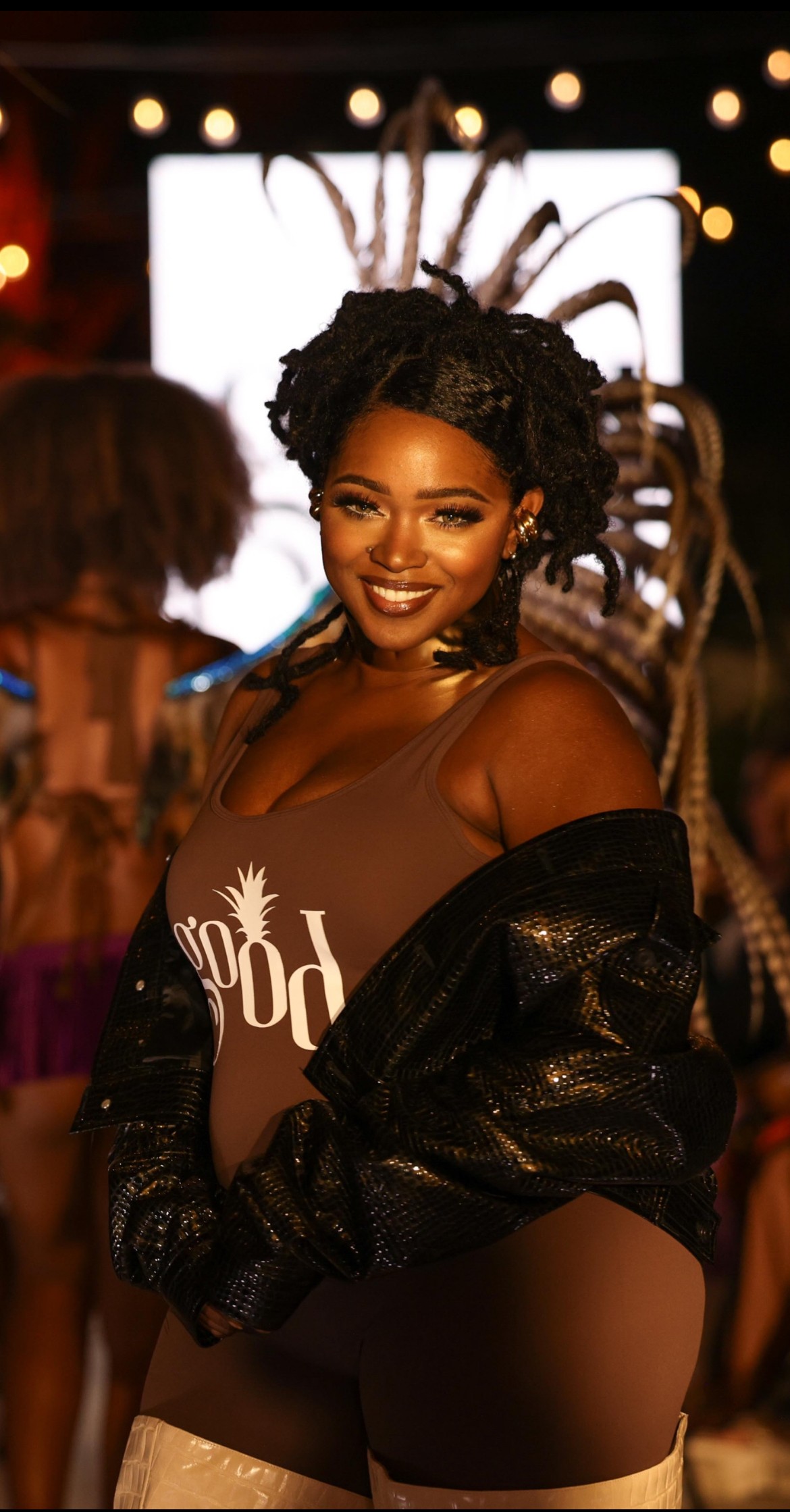 Smiling woman with styled hair, wearing a brown top and black jacket, at a festive event with blurred background lights.