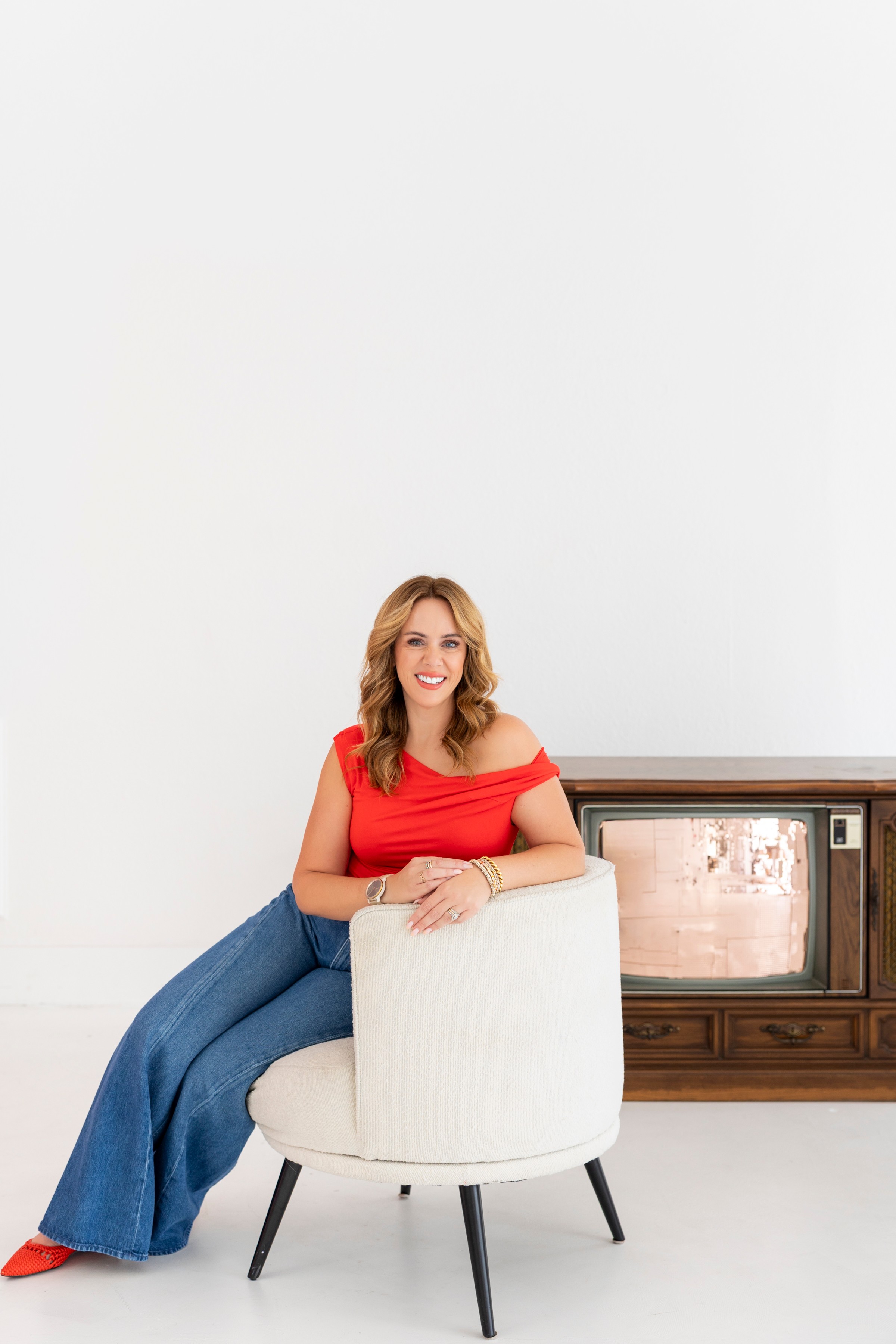 Woman with red top and blue pants sitting on a white chair, smiling, in a room with a white wall and wooden cabinet.